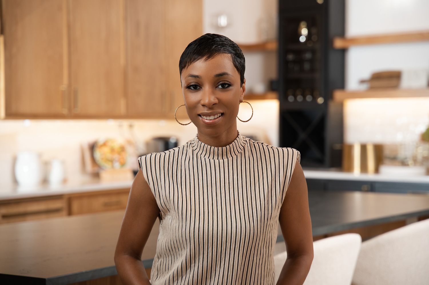 A person wearing a sleeveless striped top smiles warmly in a modern kitchen setting with wooden cabinets.