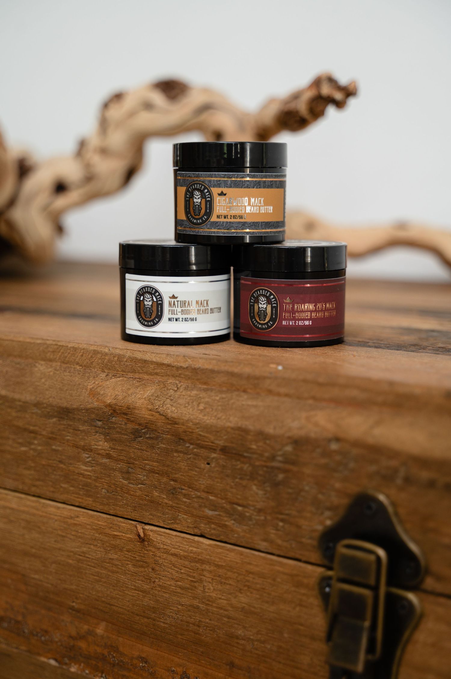 Two black cosmetic jars with brown labels sit on a rustic wooden surface with a vintage metal latch in the background.