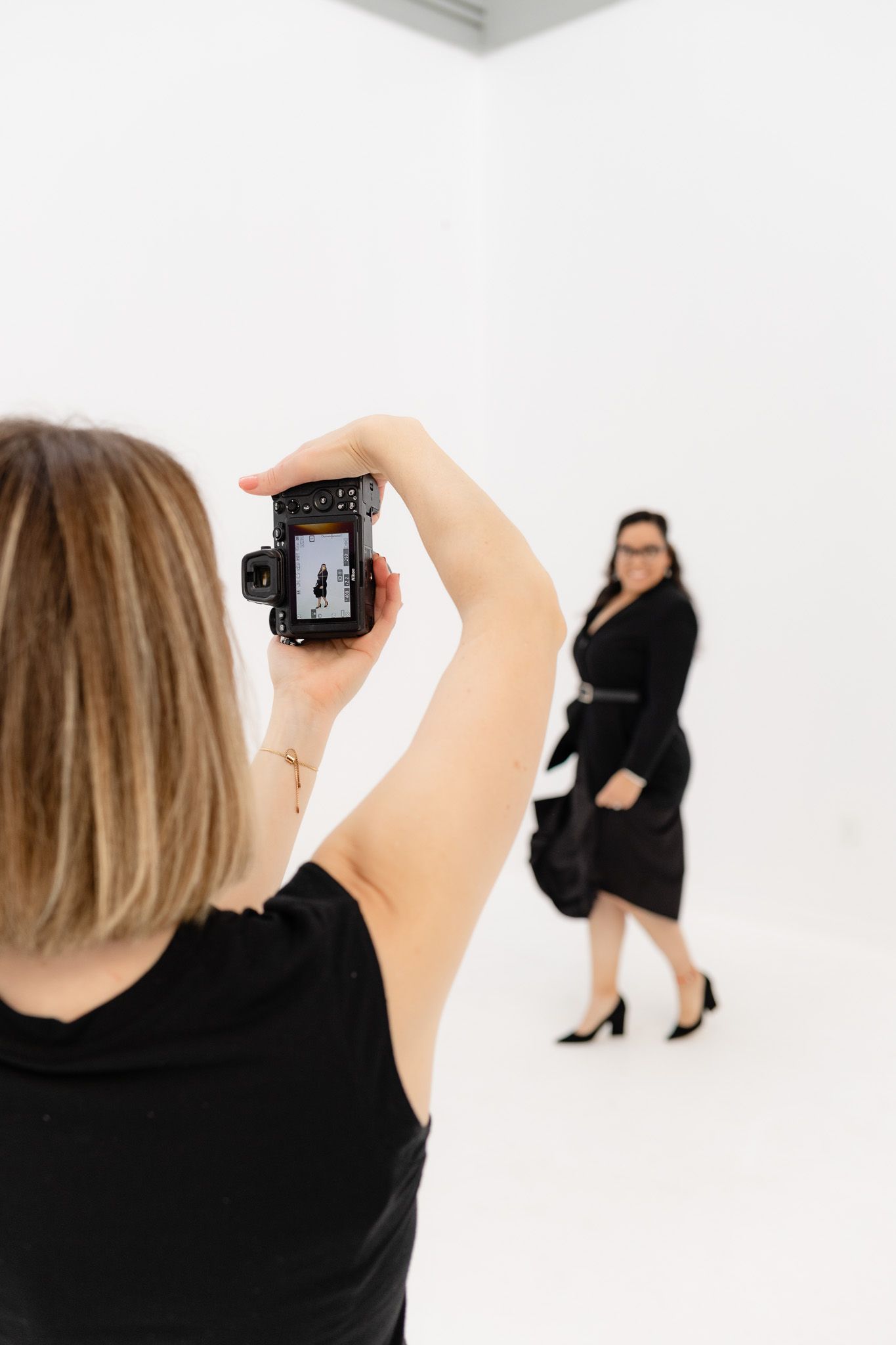 A photographer in black takes pictures of a model in a white studio setting during a professional photoshoot.
