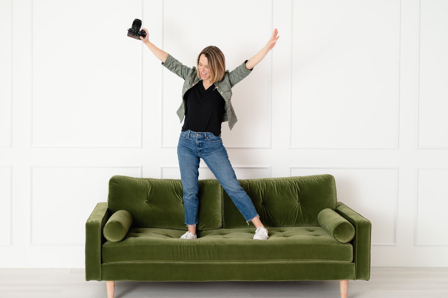 Person in casual attire demonstrates playful poses on a green velvet sofa against a white wall.