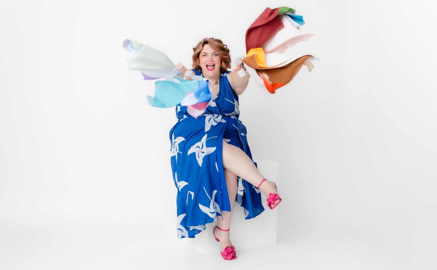 Person wearing a blue and white dress leaps joyfully against white background with colorful fabric flowing around them.