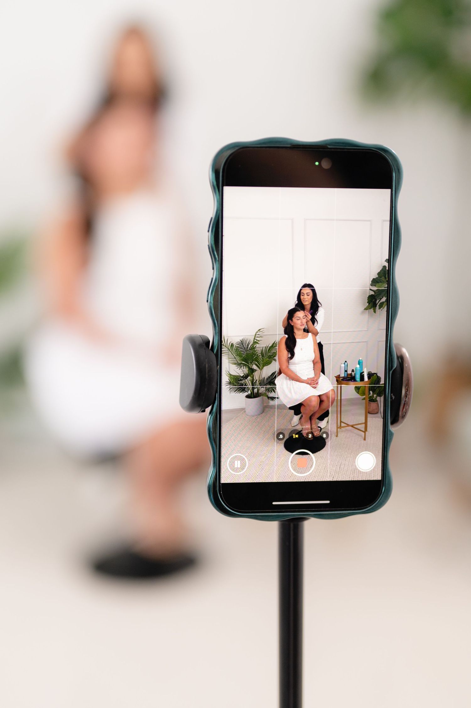 Smartphone mounted on stand displaying photo shoot setup with white backdrop and indoor plants in studio setting.