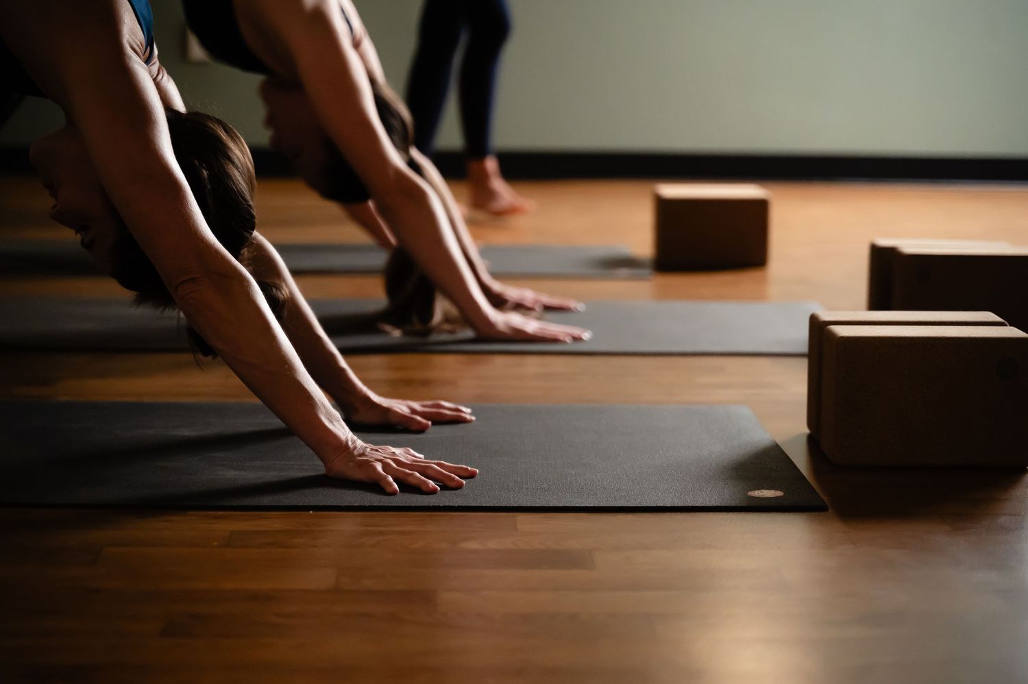 Hands press on yoga blocks during a practice session on a wooden floor in a dimly lit studio space.