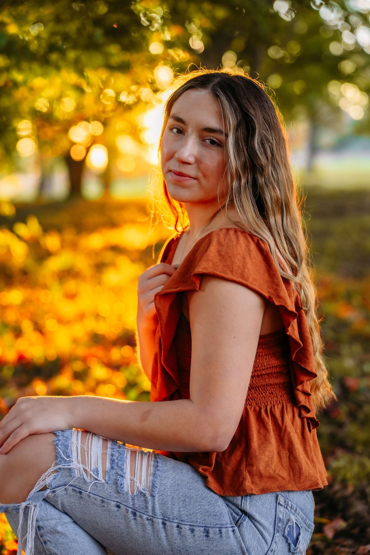 Person in orange top and jeans poses outdoors during golden hour with autumn foliage in background.