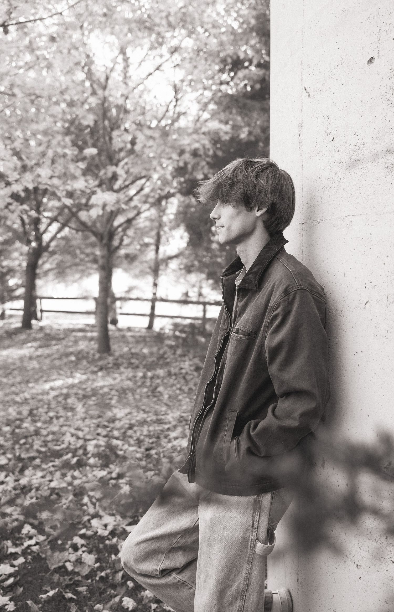 A black and white photo shows someone leaning against a wall in an autumn scene with fallen leaves on the ground.