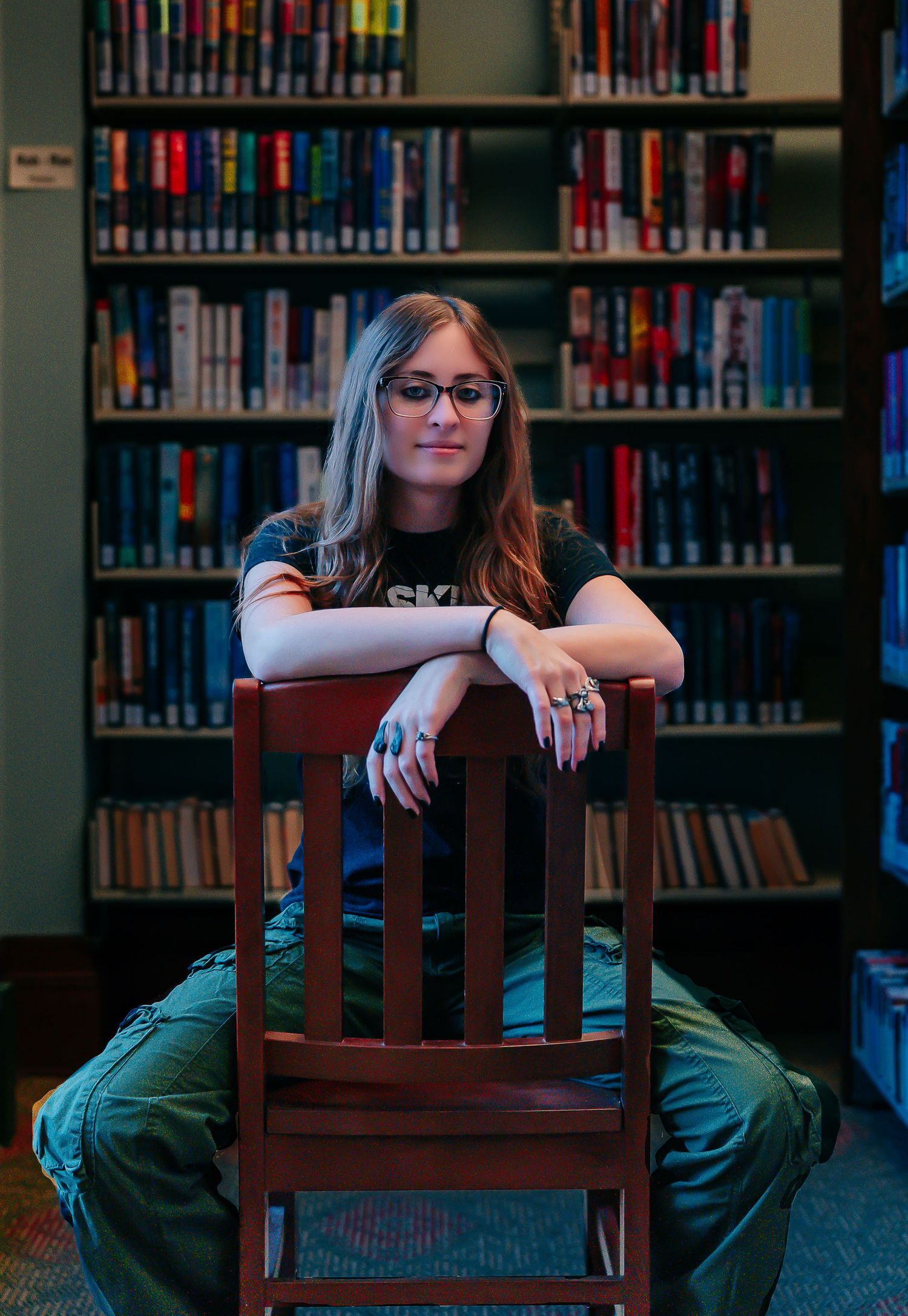 A person in glasses sits in a wooden chair at a library with bookshelves visible in the background.