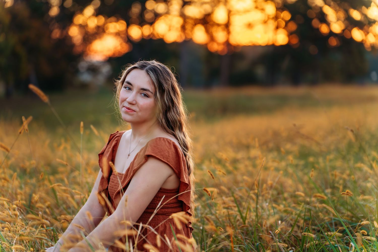 Woman in a rust-colored dress poses in a golden field at sunset during a dreamy autumn photoshoot.