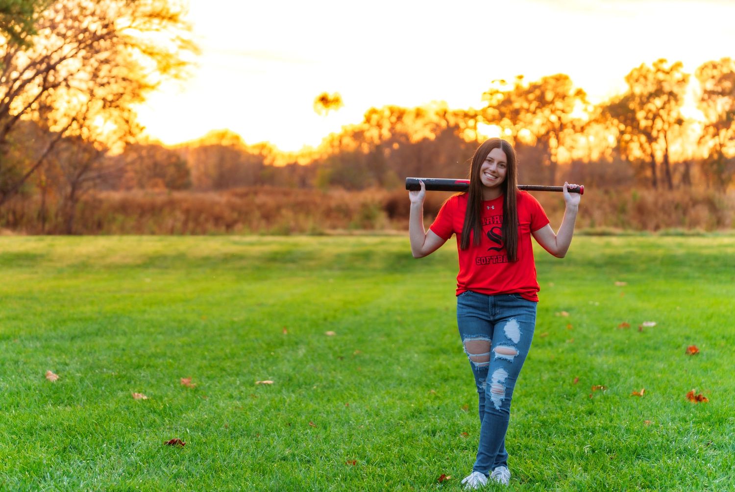 A person in a red shirt and jeans stands in a vibrant green field at sunset during autumn.