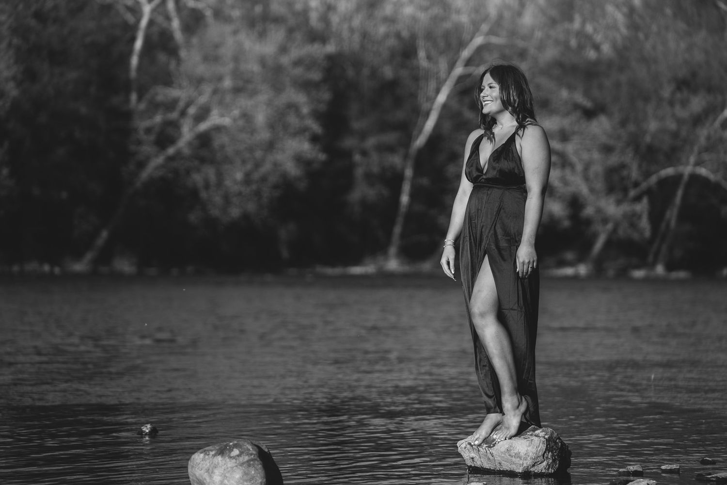 A woman in a black dress wades through a tranquil lake surrounded by trees during a photo session.