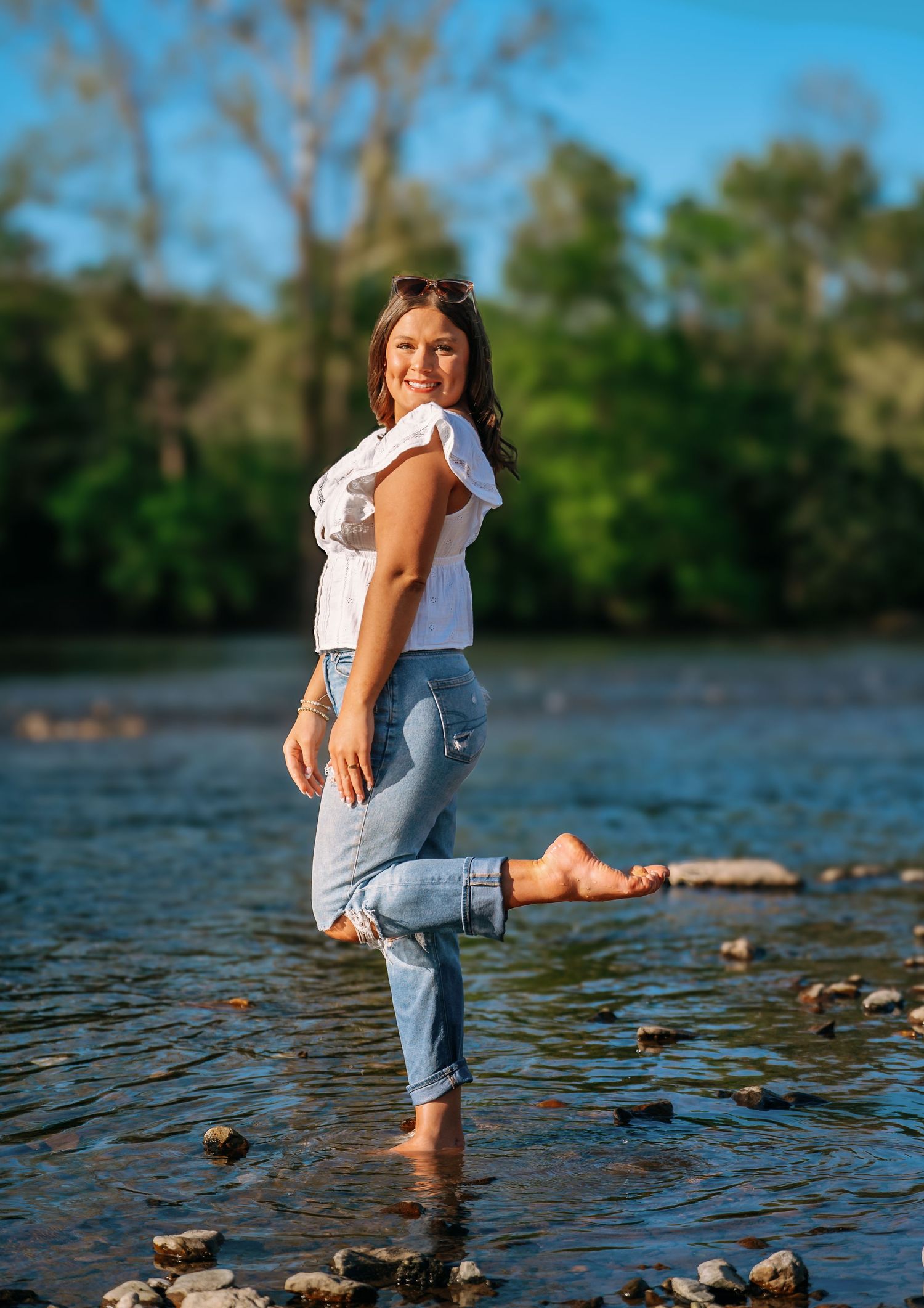 Person in jeans sitting by a river's edge with trees in the background.