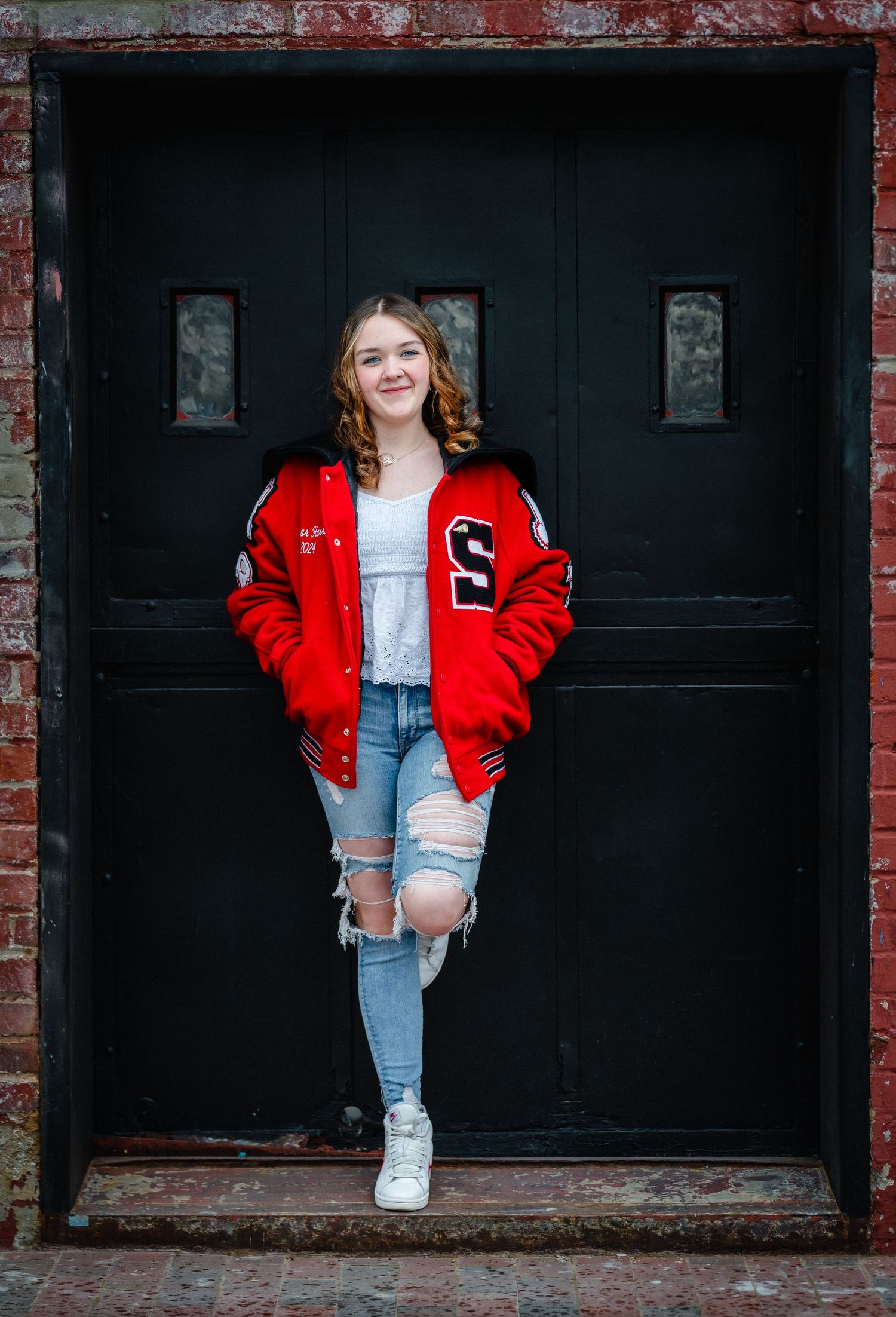 Casual portrait against brick wall wearing red varsity jacket, distressed jeans and white sneakers.