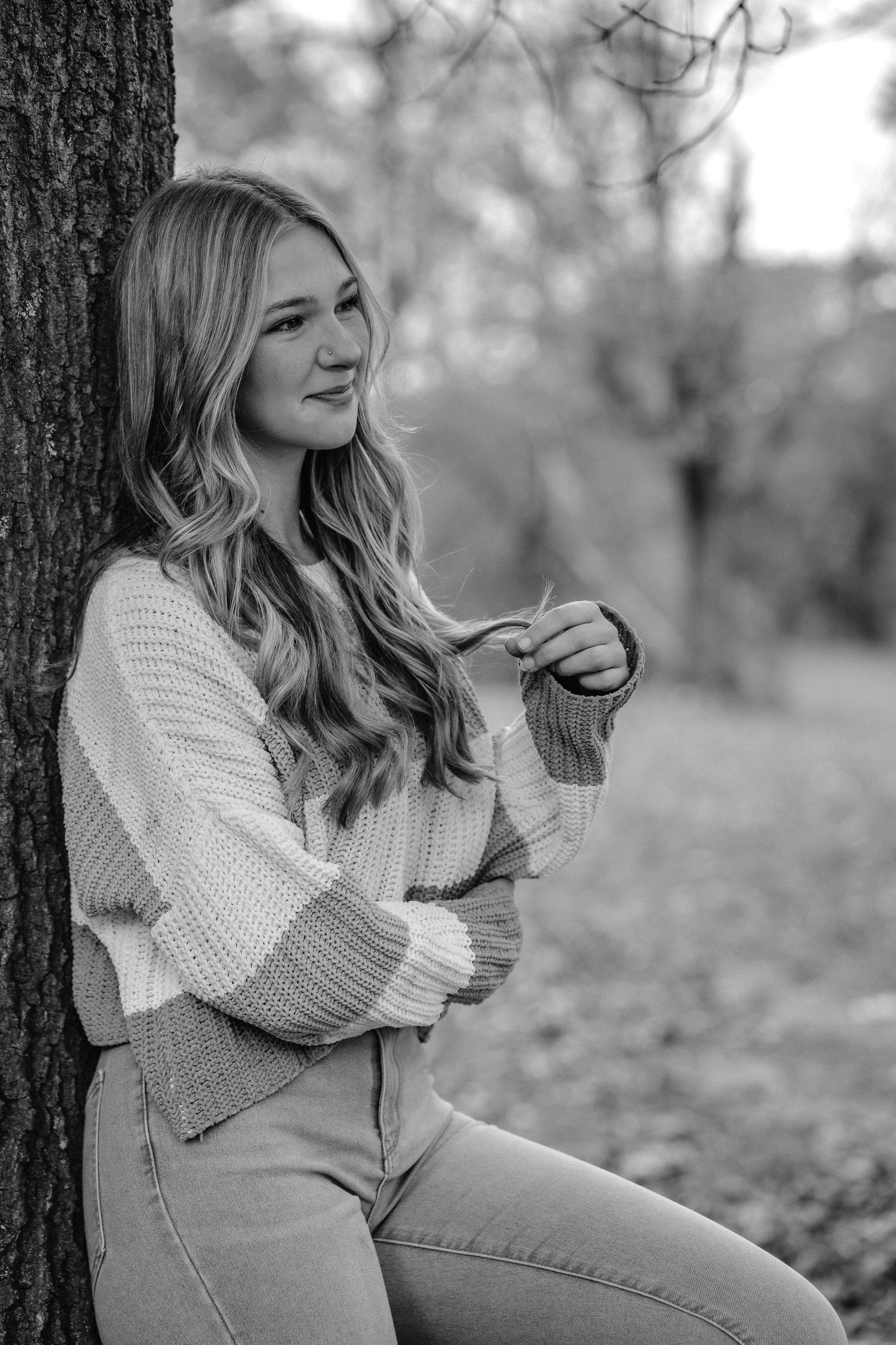 Black and white portrait series showing someone in a cozy sweater sitting against a tree trunk in an autumn setting.