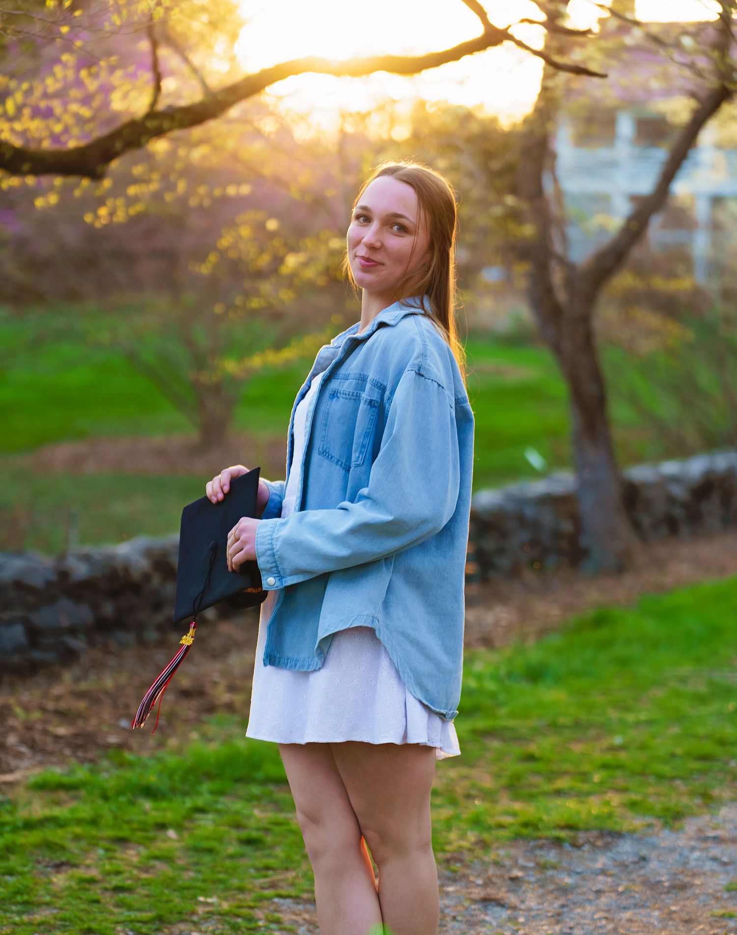 A woman in a denim jacket poses in a park during golden hour with trees and sunlight in the background.