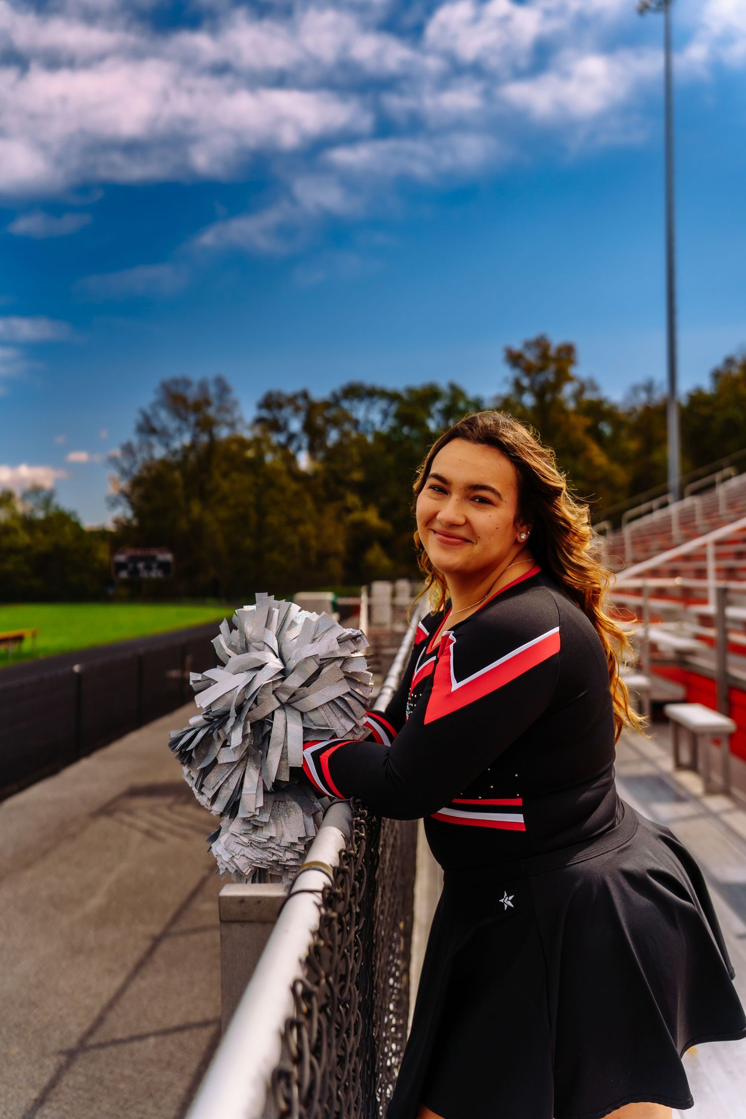 A cheerleader in black and red uniform poses with pom poms against bleachers and blue sky on a sunny day.