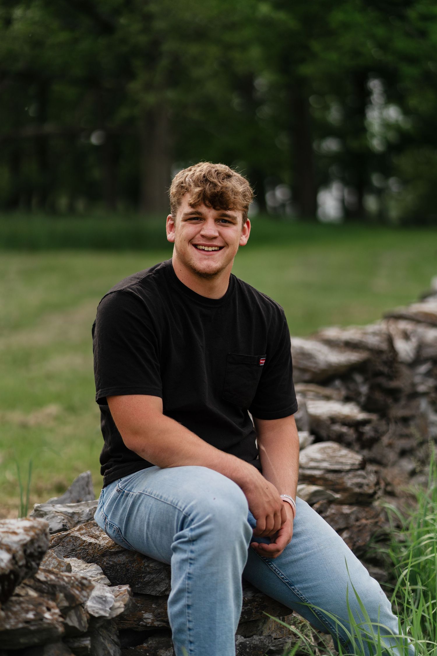 A person in a black t-shirt and jeans sits casually on a rustic stone wall surrounded by green grass.