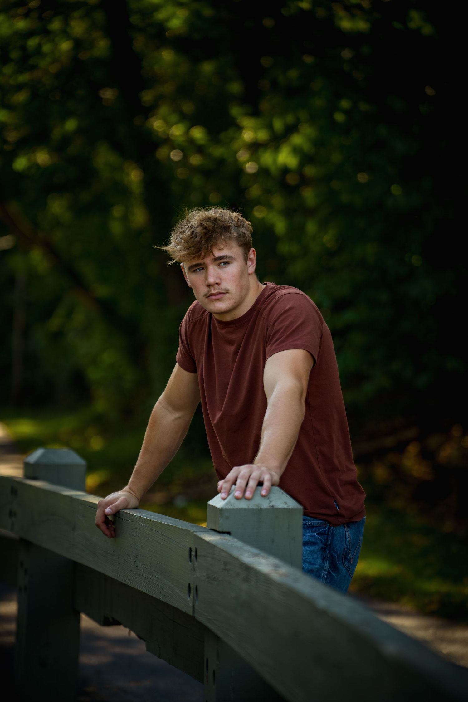 A person in a burgundy t-shirt leans on a railing outdoors in a park setting with natural lighting.