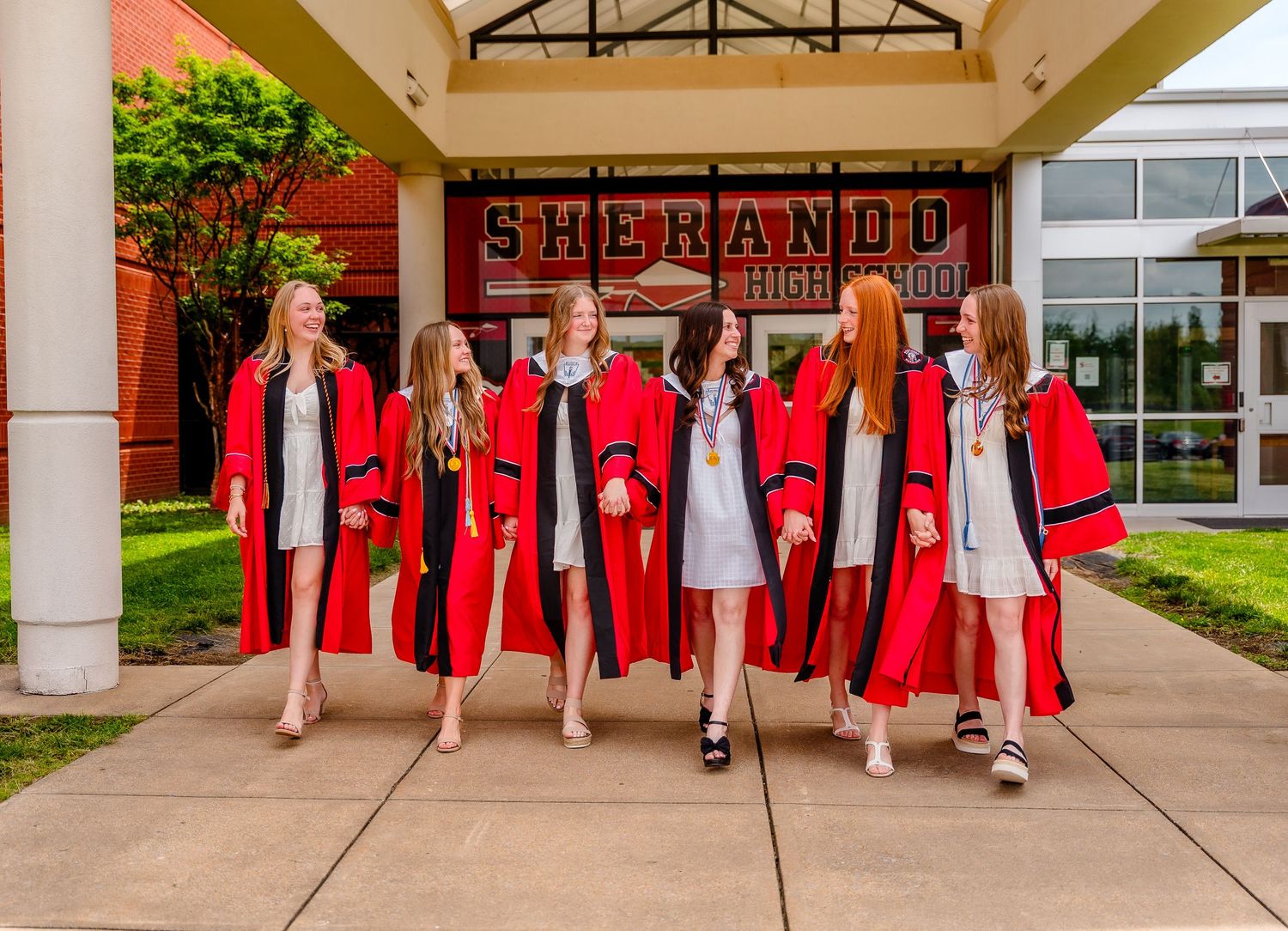 Row of graduates in red caps and gowns pose together outside school building.
