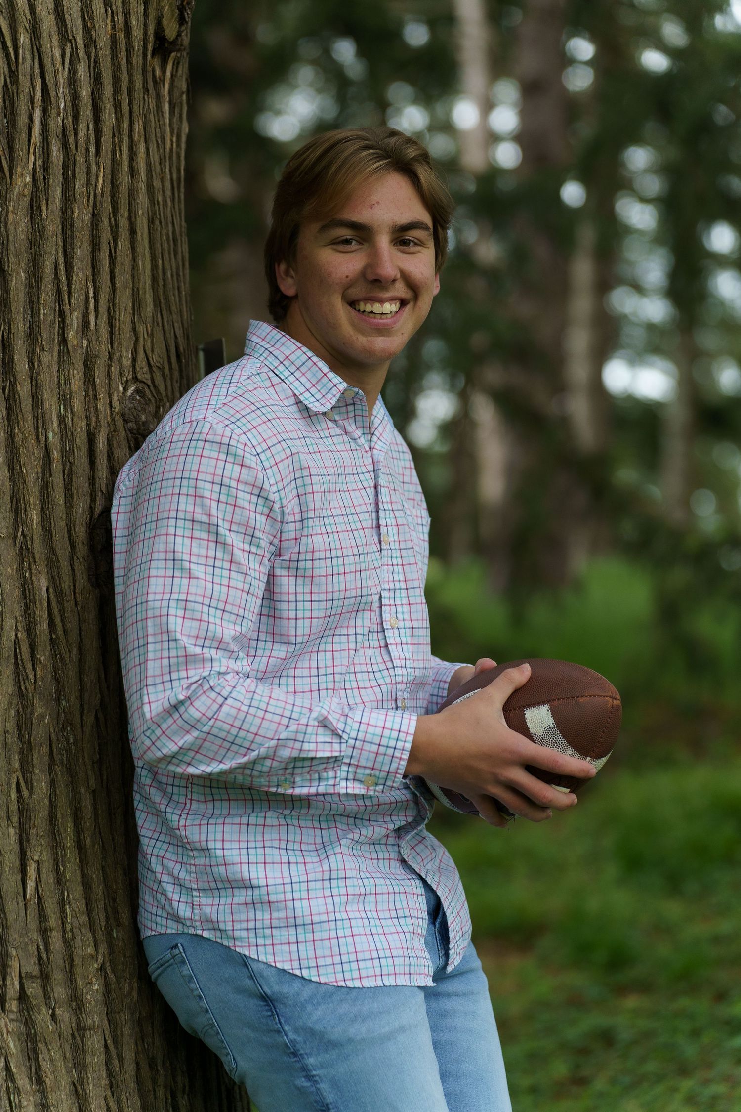 Person in plaid button-down shirt and jeans sits casually against tree trunk in outdoor wooded setting.
