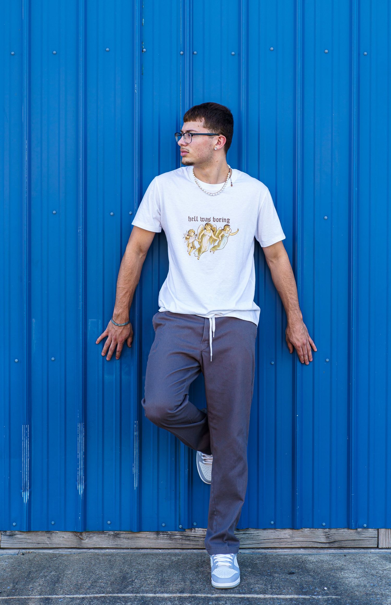 Someone wearing glasses, a white graphic t-shirt and grey sweatpants poses casually against a bright blue metal wall.