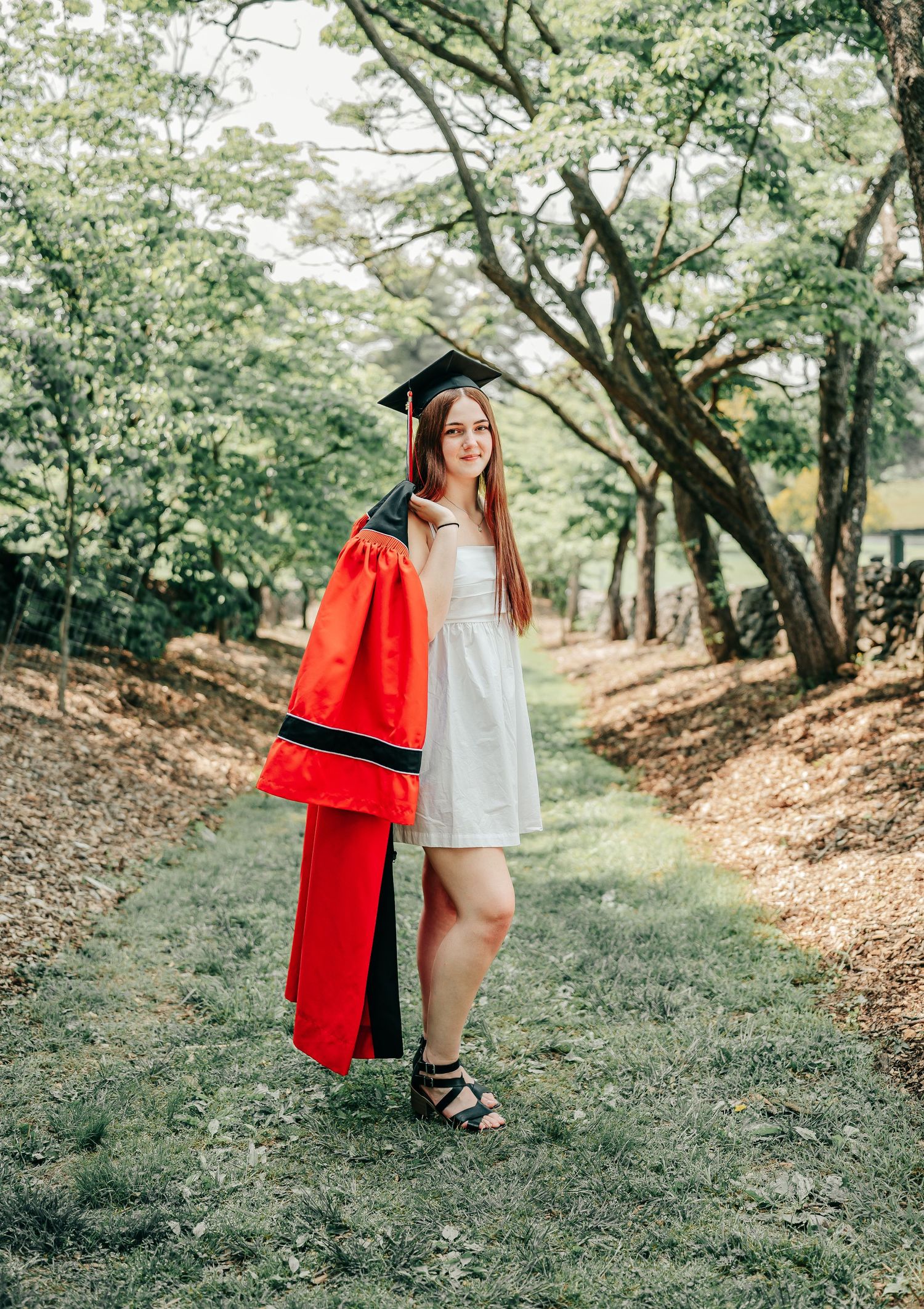 High school graduate girl in white gown holding her red graduation gown along a tree-lined path during a sunny outdoor photoshoot.