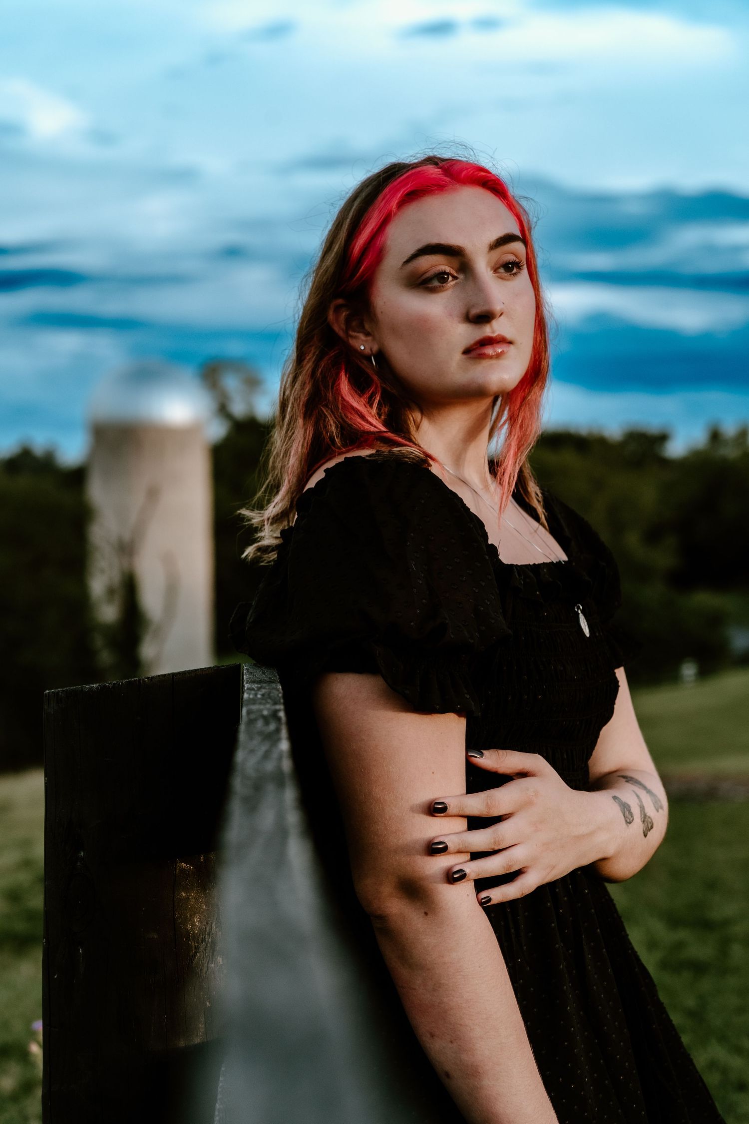 Portrait of a woman with vibrant red hair in a black dress leaning against a fence post at sunset.