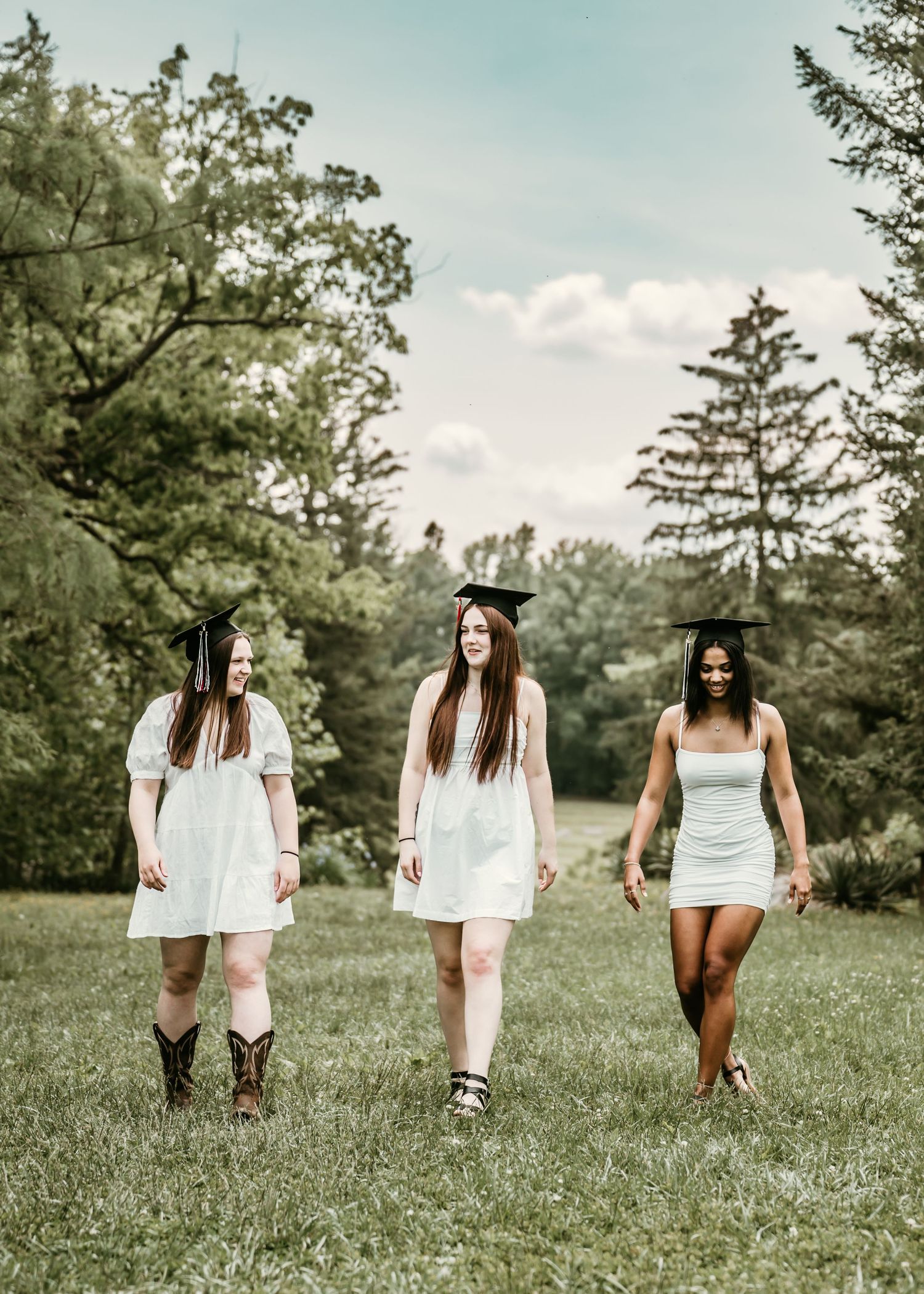 Group of high school senior girls walking towards the camera across a grassy field wearing white dresses and boots on a sunny day.