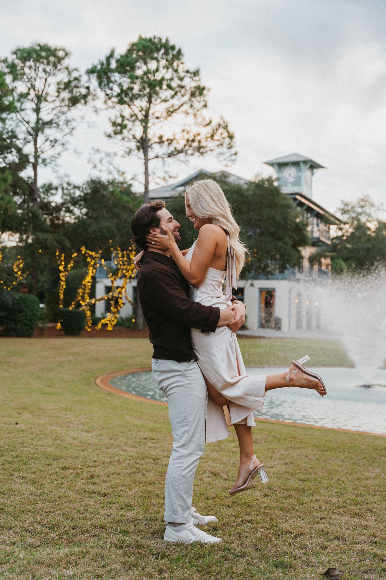 A couple shares a romantic embrace by a fountain with twinkling lights in the background.