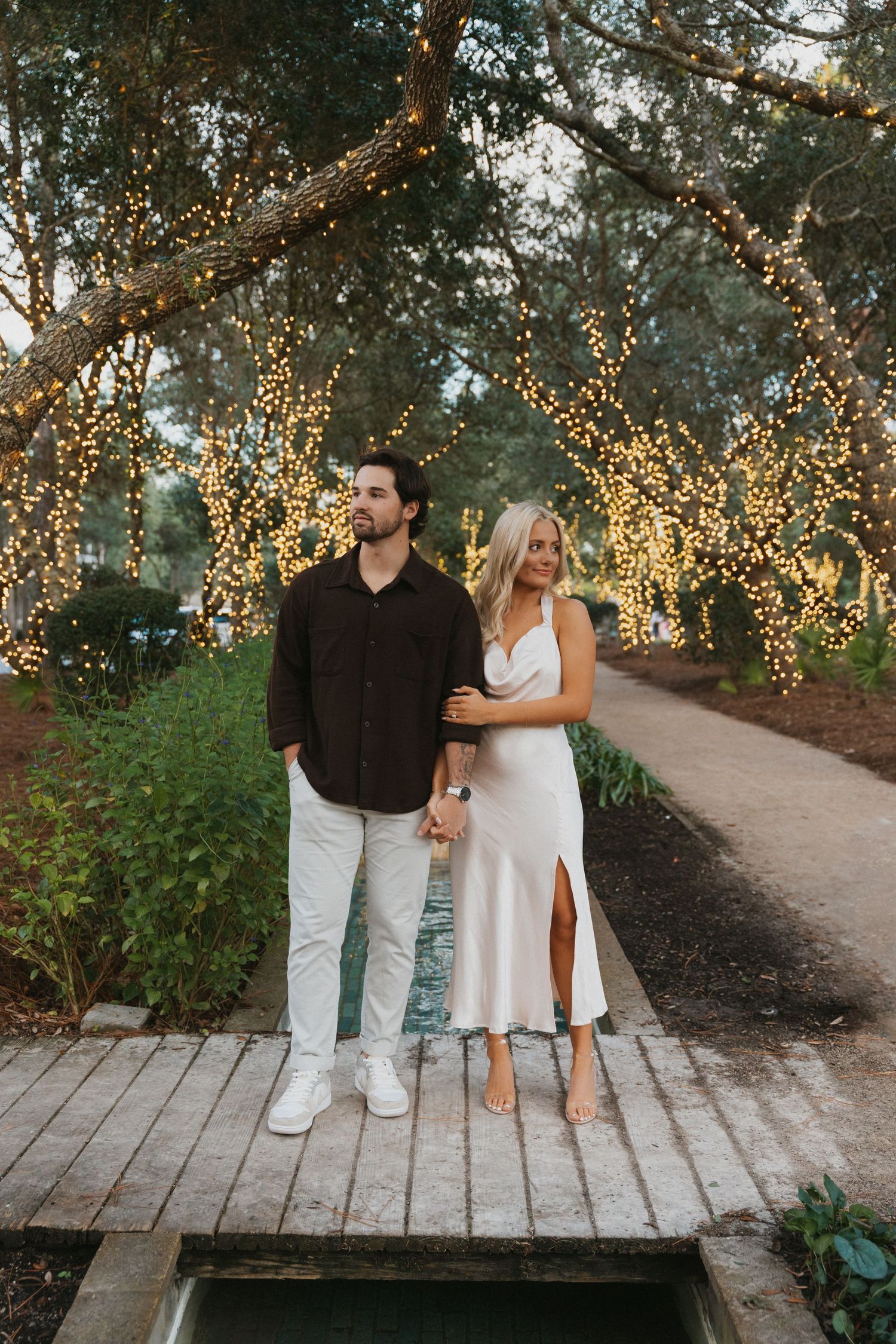 A couple poses on a wooden bridge with illuminated trees creating a magical evening atmosphere.