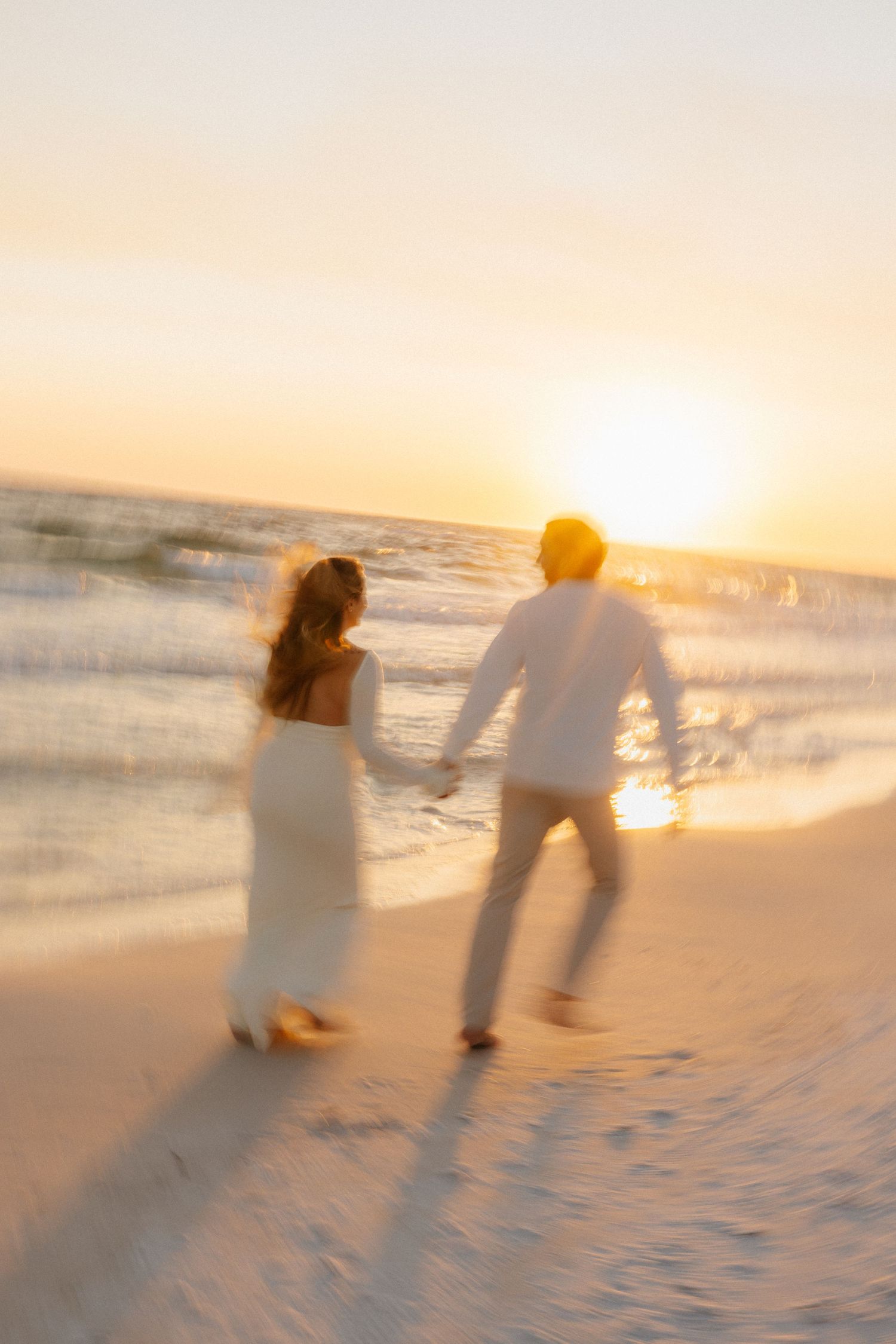 A couple in white attire walks hand in hand along a beach during golden hour sunset, creating a dreamy silhouette.