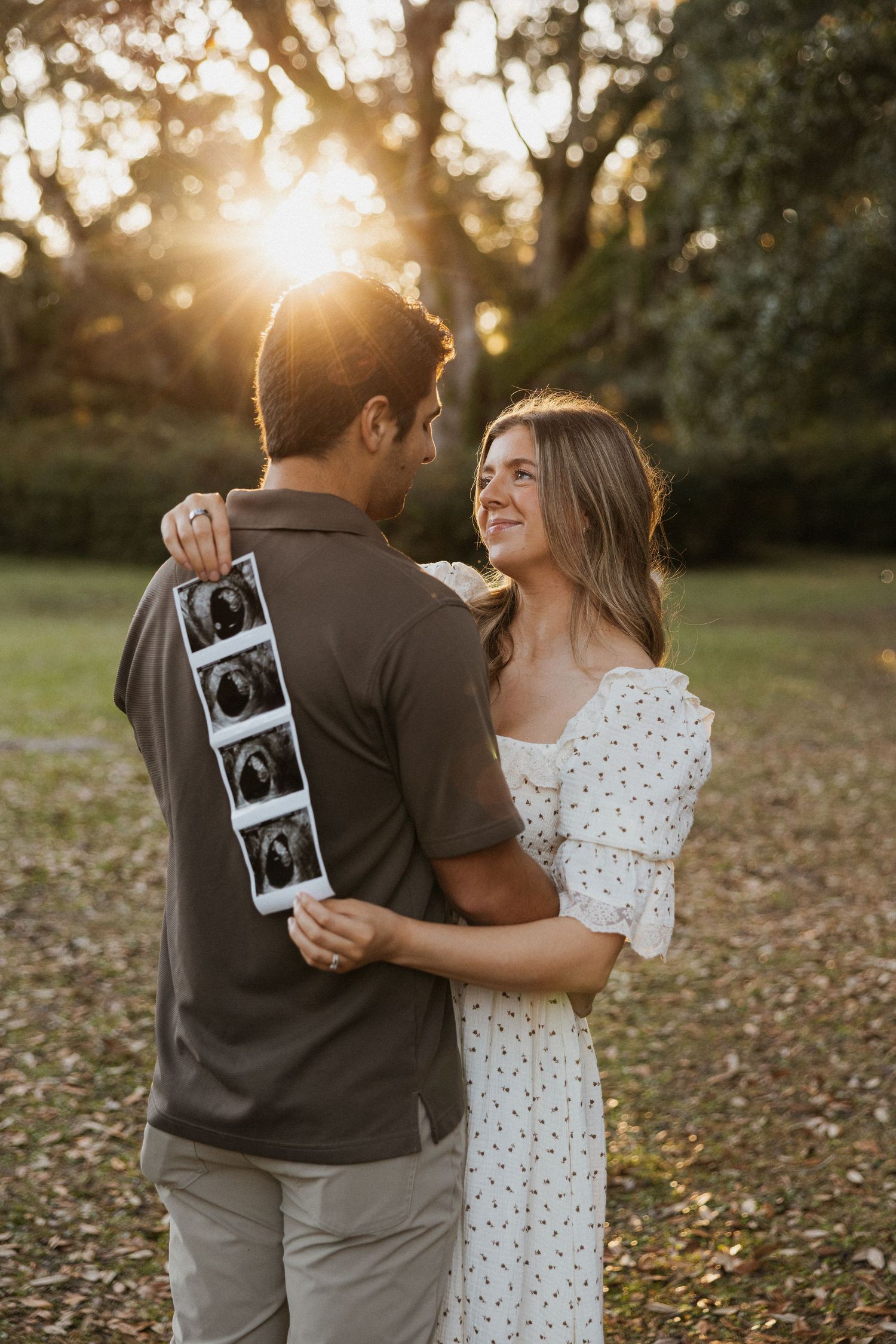 Couple in a park at sunset holding up ultrasound photos while sharing a joyful moment together.