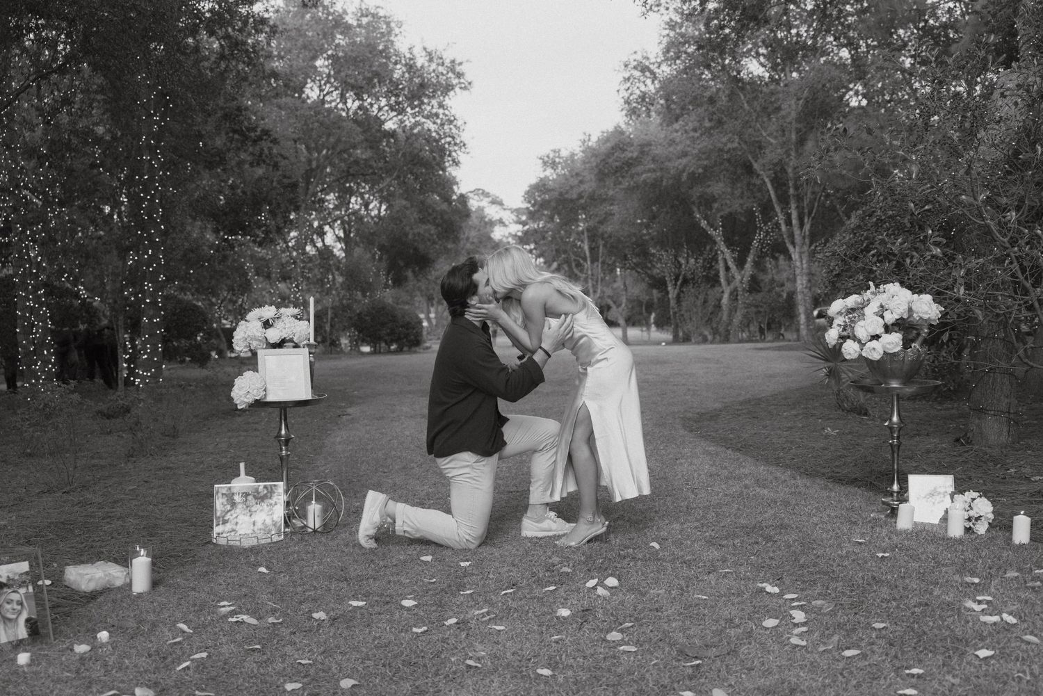 A black and white photo capturing a proposal moment in a garden setting with scattered decorations.