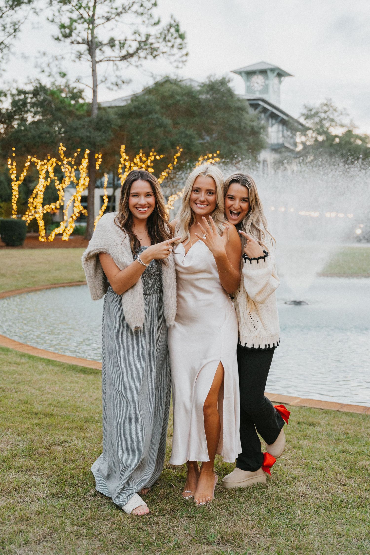 Three friends celebrate together near a fountain with festive lights in the background.
