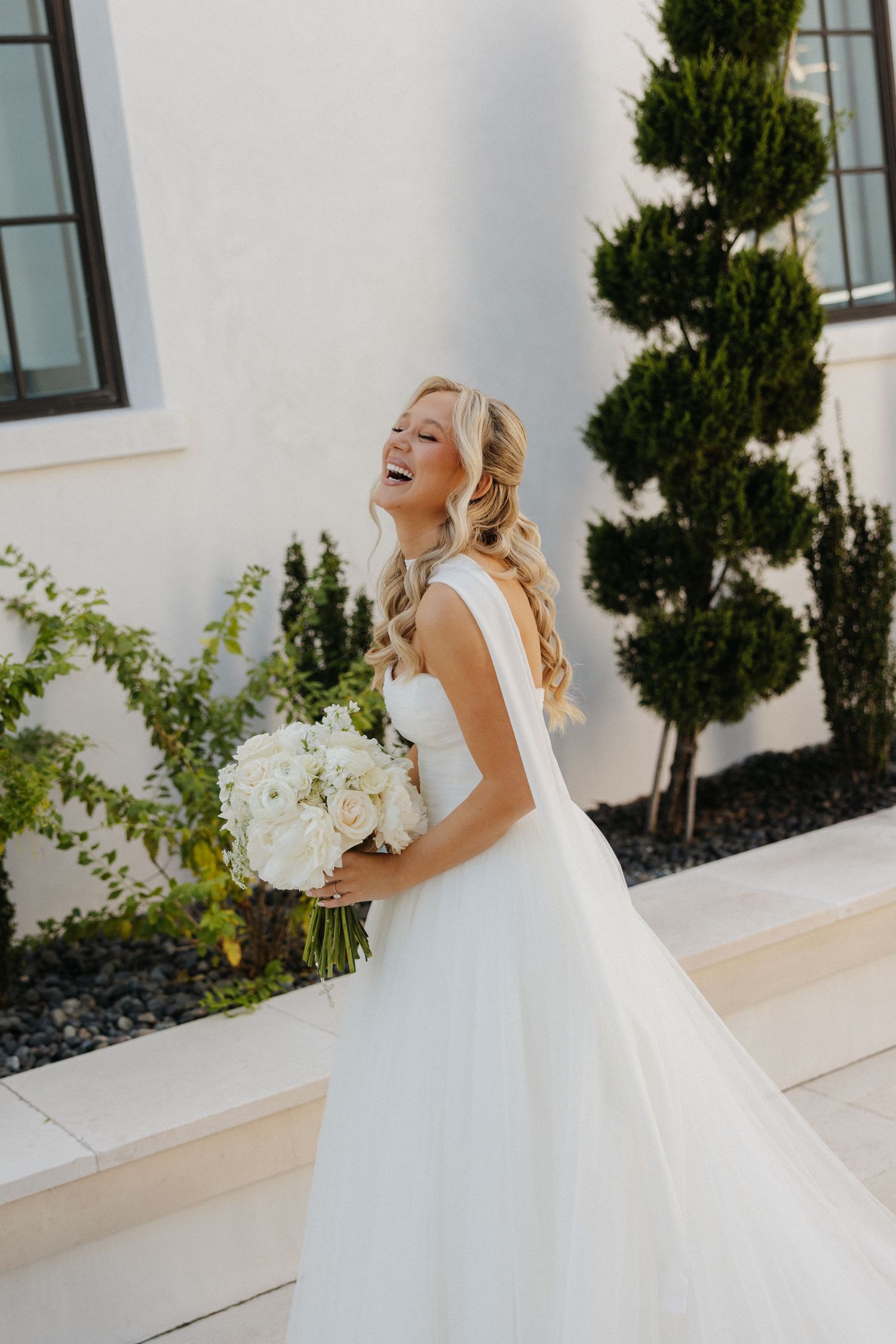 A joyful bride in a white ballgown twirls and laughs on a bright sunny day outside a white building.