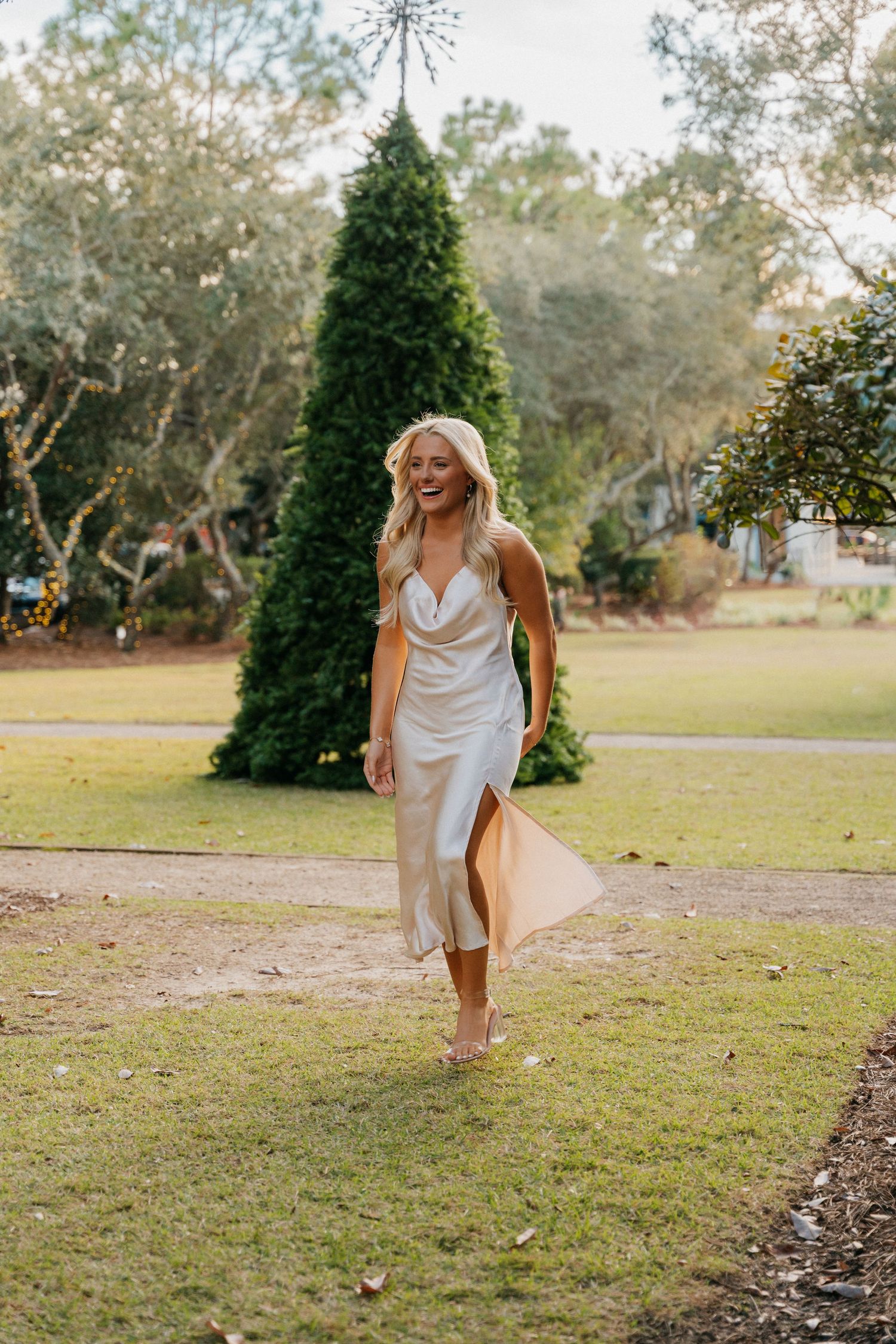 A woman in a flowing silk dress spins joyfully in a garden with a large topiary tree.