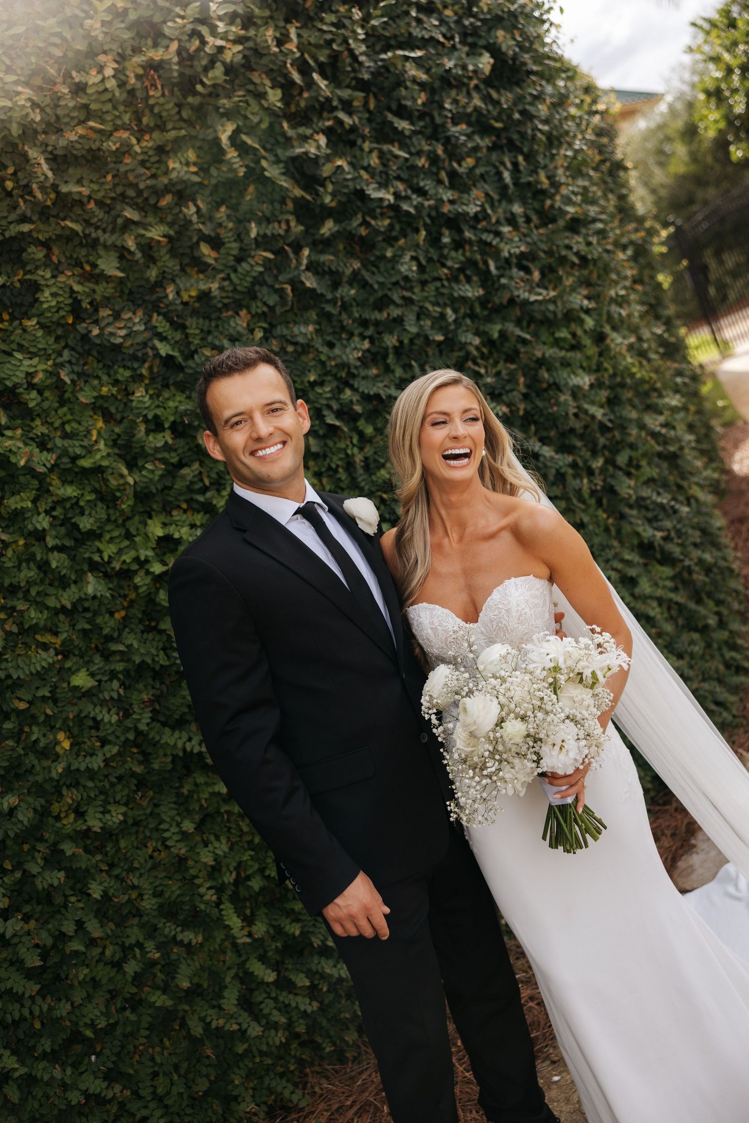 Joyful wedding couple laughing together against lush green hedge backdrop in outdoor garden setting.