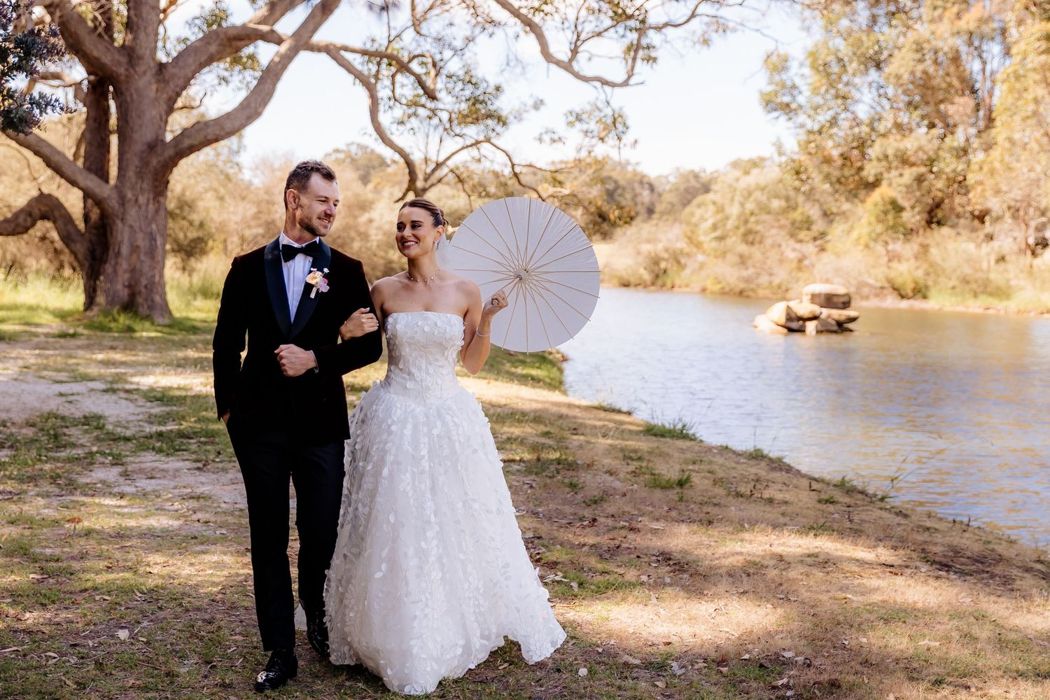 A couple in formal wedding attire poses together by a scenic riverside on a sunny autumn day.