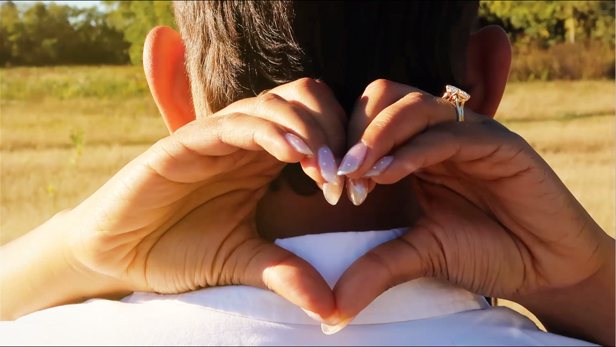 Woman making a heart with her hands behind her fiancé's back for engagement video