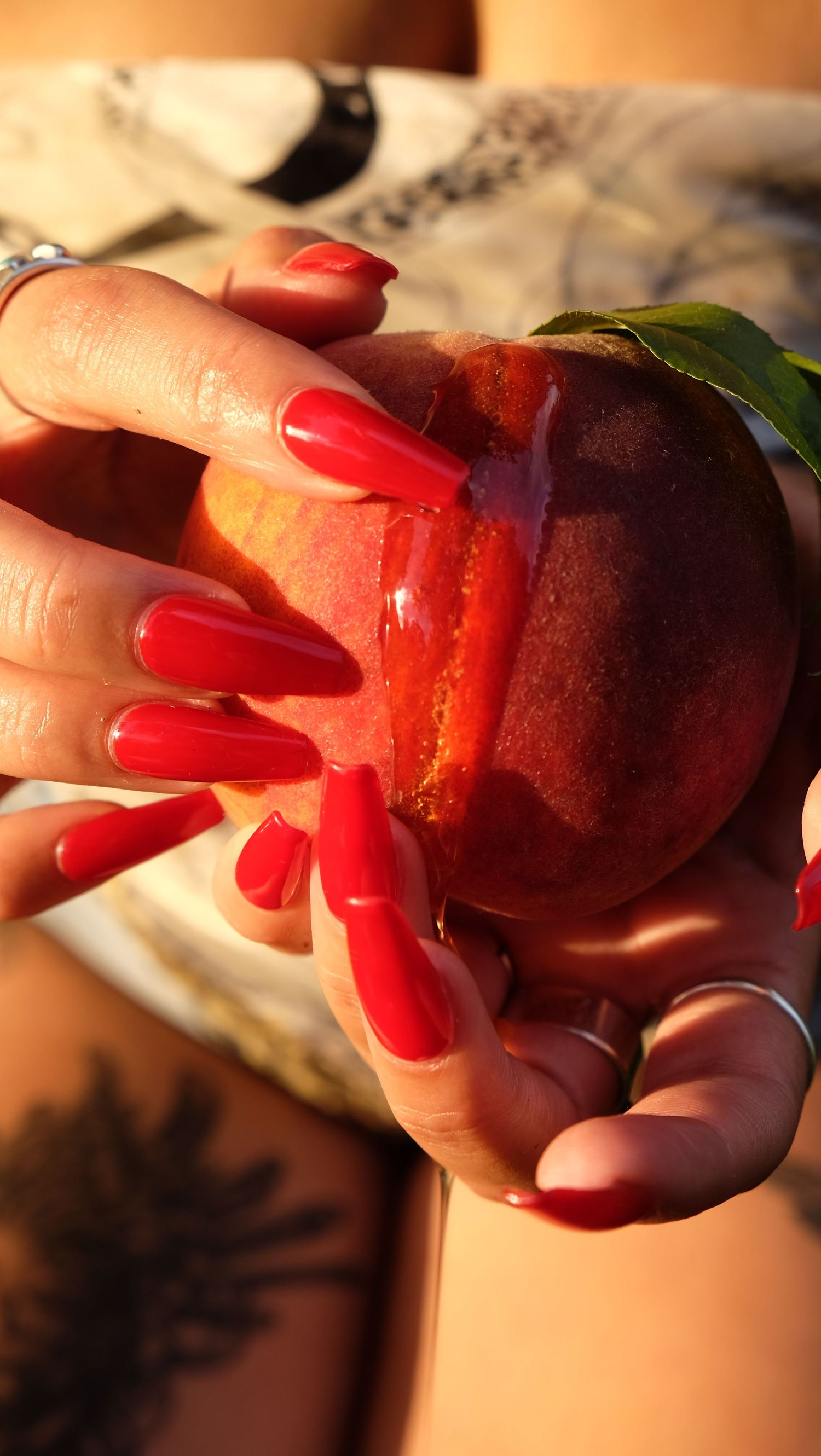 Hands with long red nails and silver rings hold a ripe peach glistening with honey in warm sunlight.