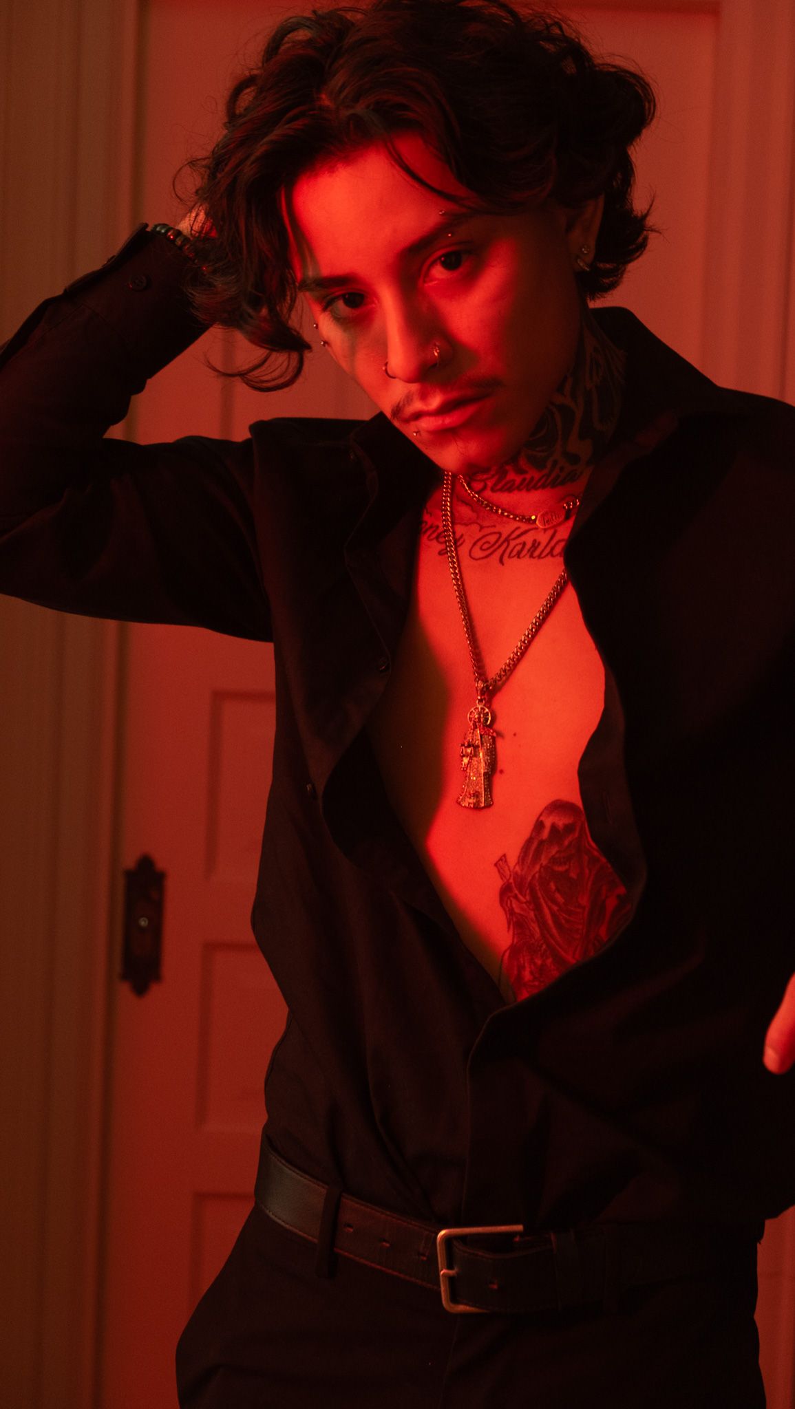 Man with wavy dark hair, face piercings and chest tattoo poses in an open black shirt bathed in red light.