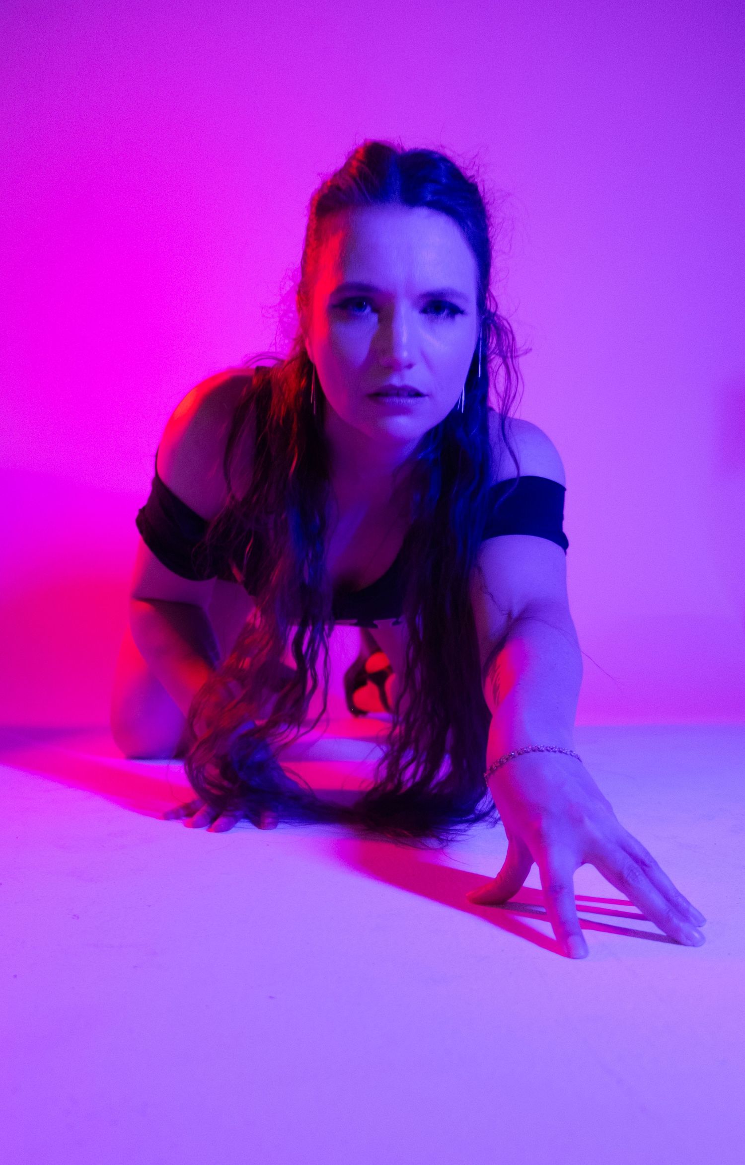 Woman with long dark hair crouching and gazing at the camera in a studio bathed in vibrant magenta and blue lighting.