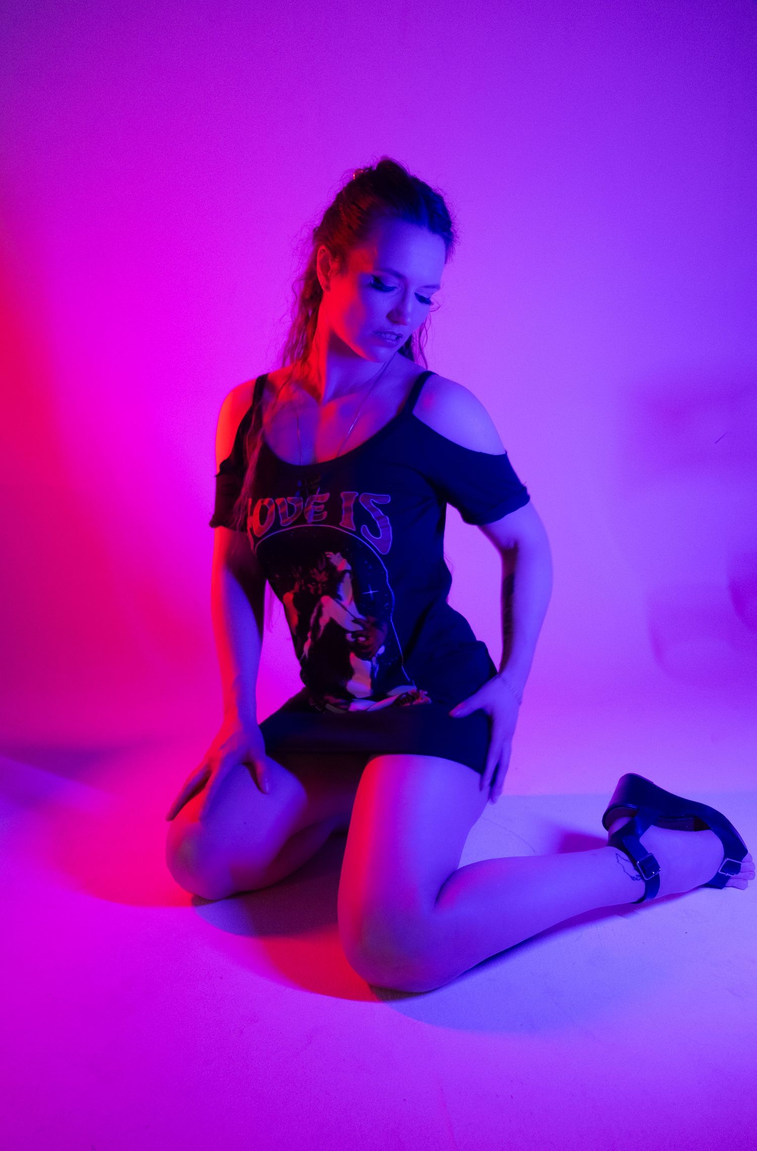 Woman in an off-shoulder graphic tee and black heels kneels in vivid pink and purple studio lighting.