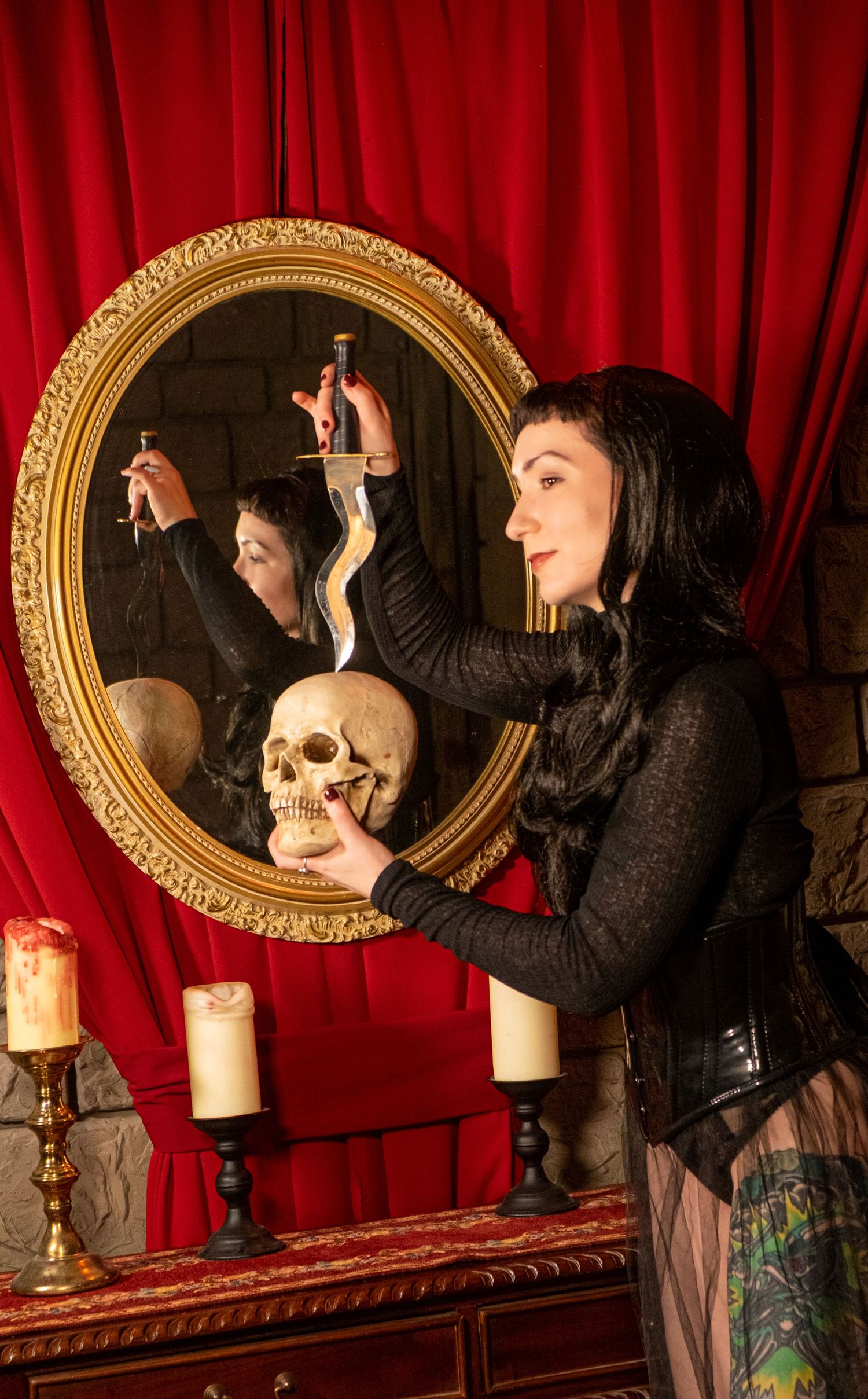 Dark-haired woman in a corset holds a dagger above a skull before an ornate gold mirror and red velvet curtain.