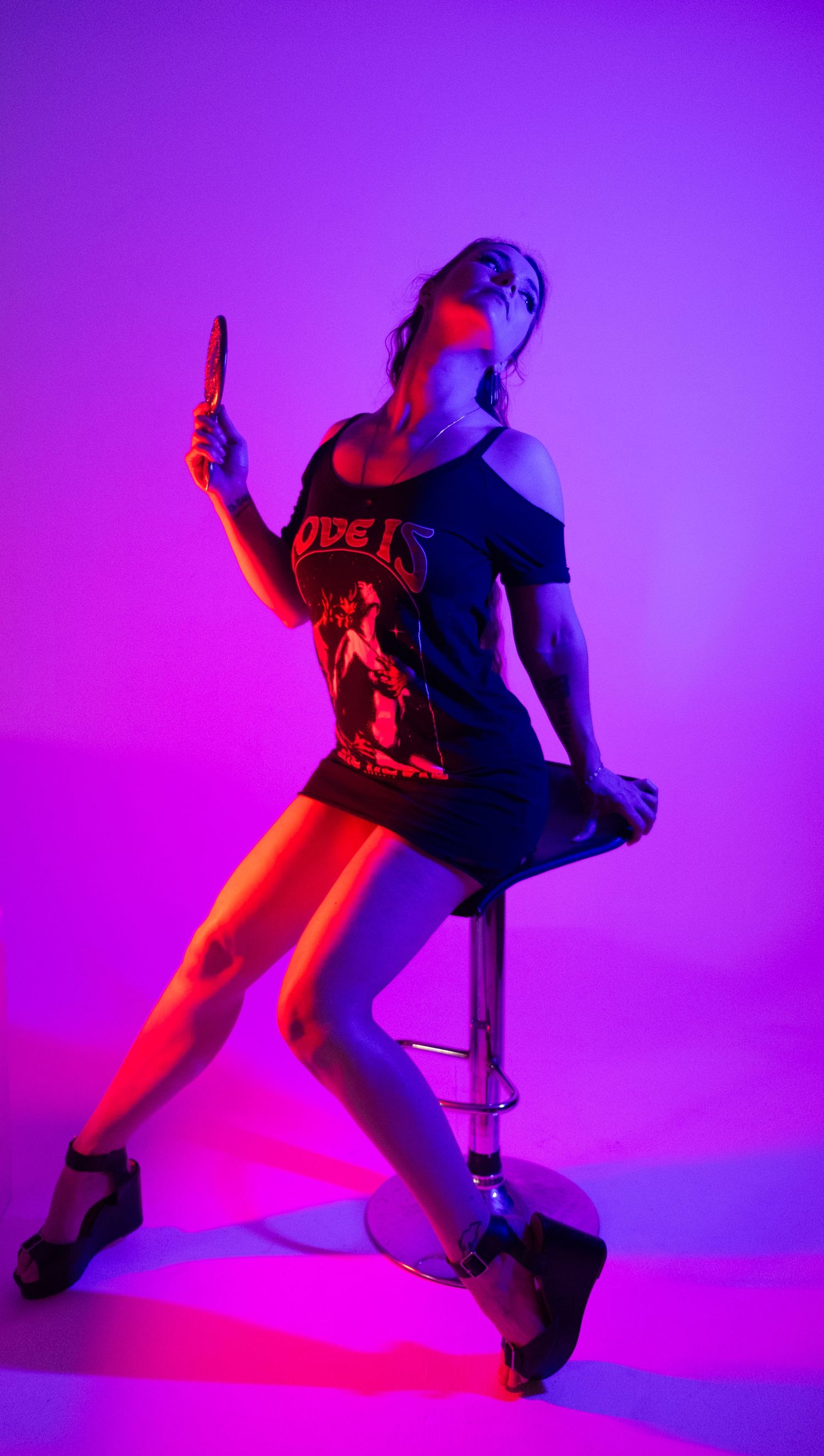 Woman in a graphic tee and platform heels sitting on a bar stool, bathed in vibrant magenta and purple studio light.