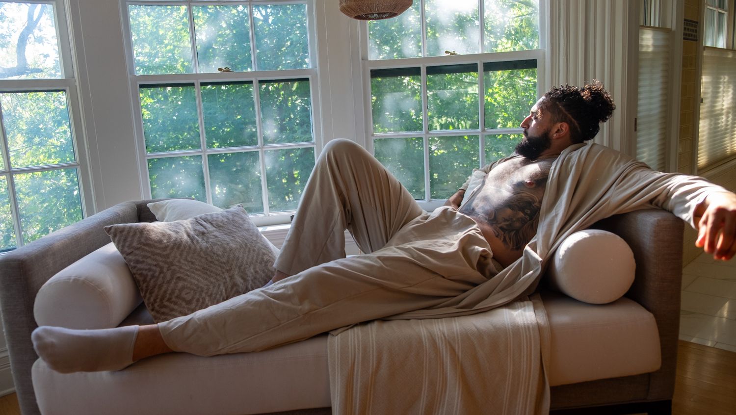 Bearded man with a bun lounges on a daybed in loose beige clothing beside sunlit windows with green trees outside.