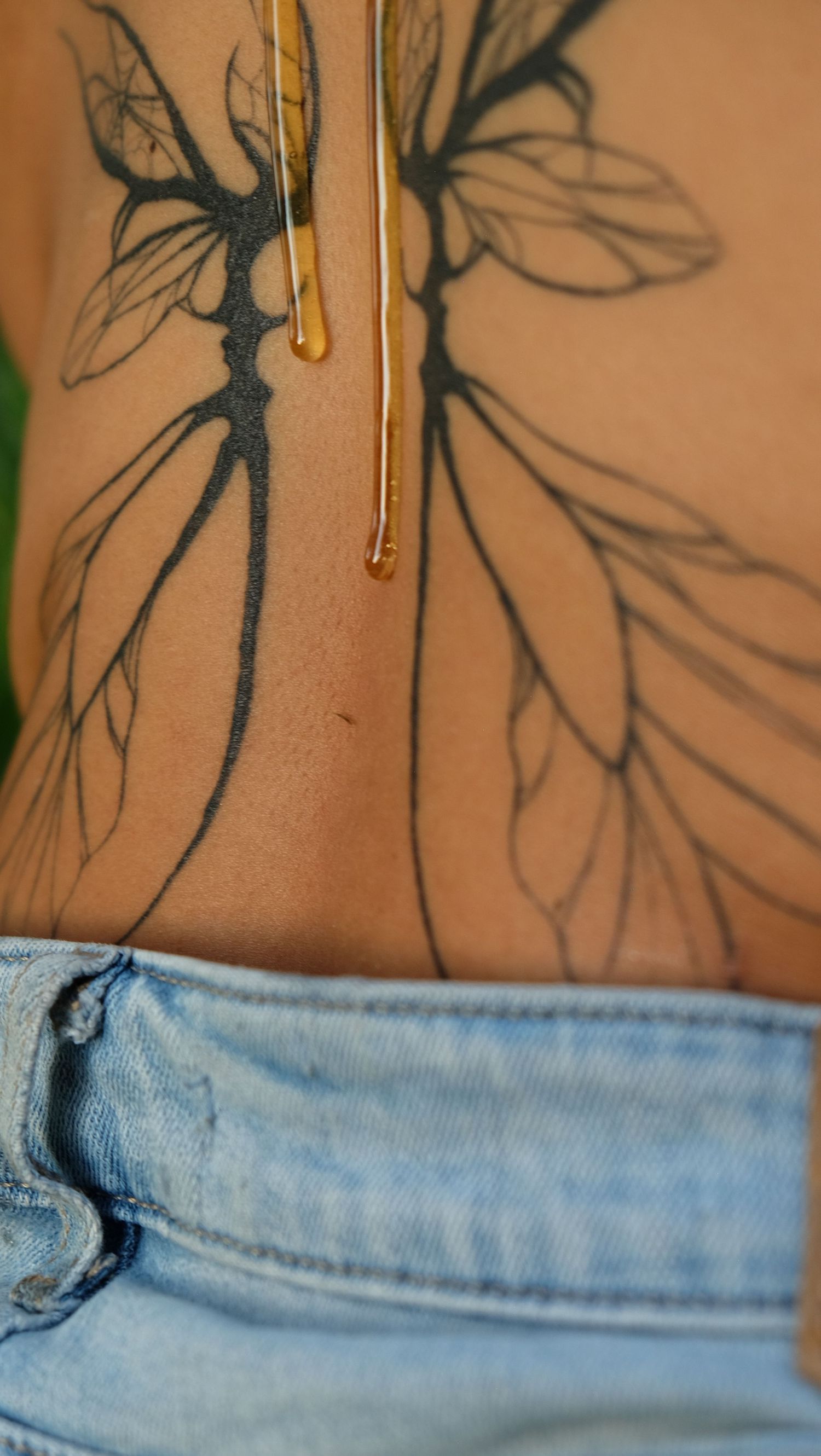 Honey drips onto a tattoo of dragonfly wings across a person's lower back above blue denim jeans.