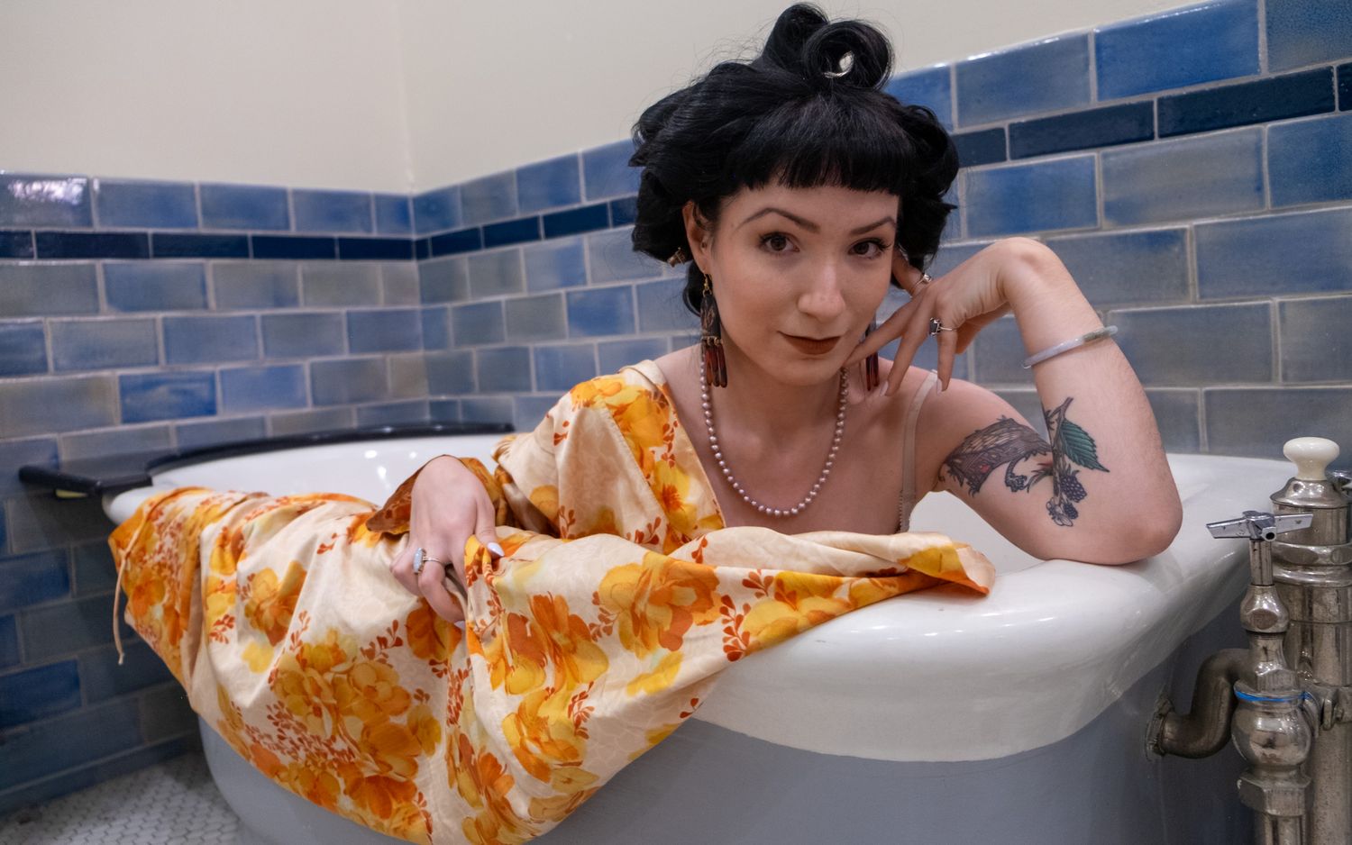 Woman with dark updo and tattooed arm reclines in a clawfoot bathtub wearing a yellow floral robe and pearl necklace.