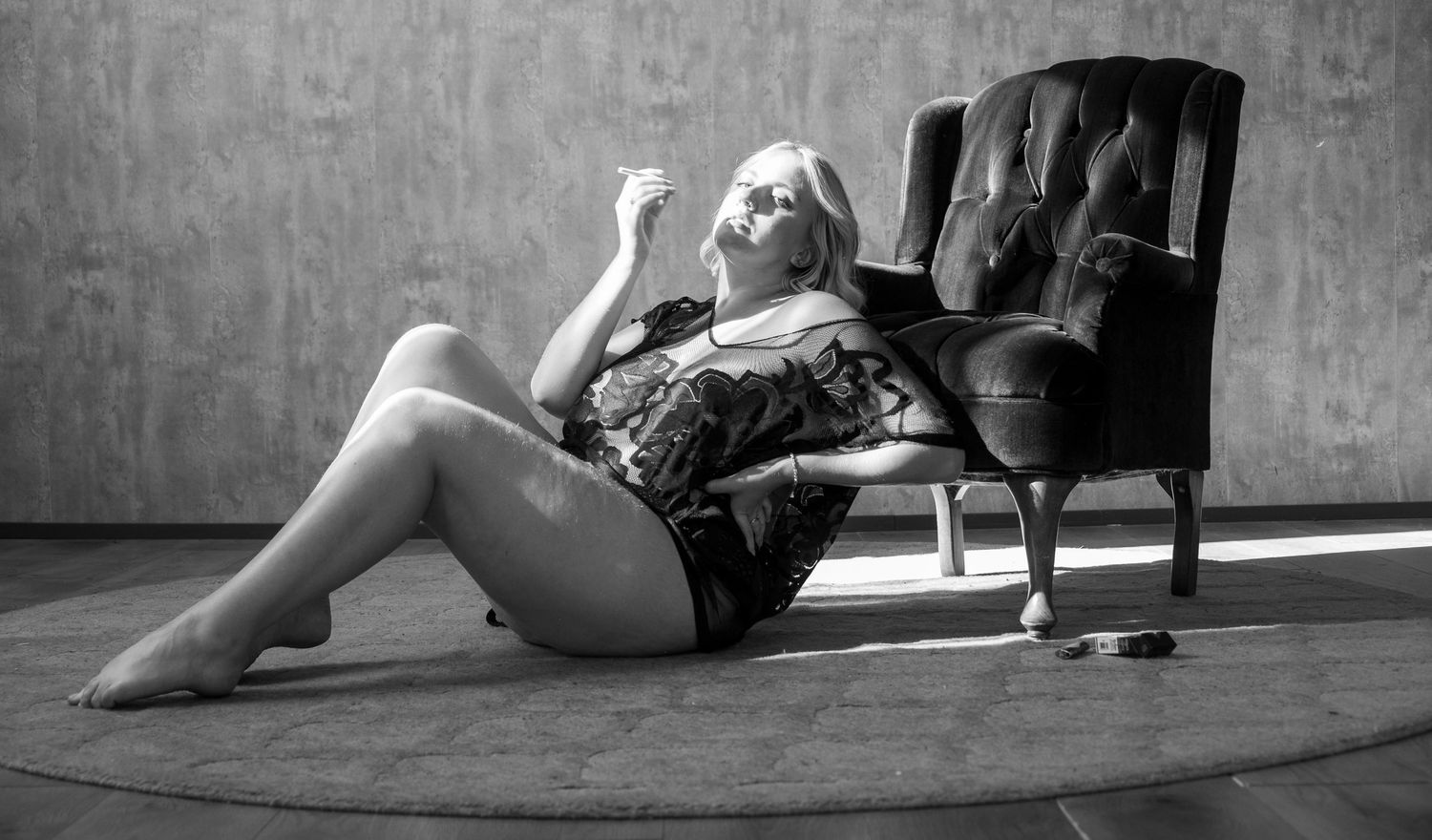 Black and white photo of a blonde woman sitting on the floor beside a tufted armchair on a round rug in soft window light.
