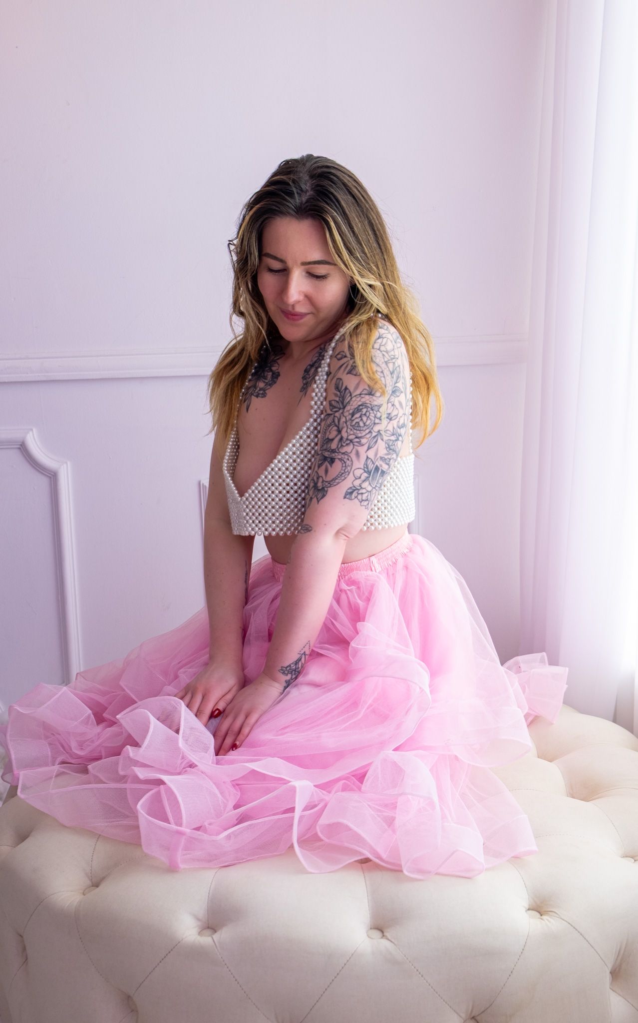 Tattooed woman in a pearl-beaded top and pink tulle skirt sits on a cream tufted ottoman, looking down in a pastel studio.