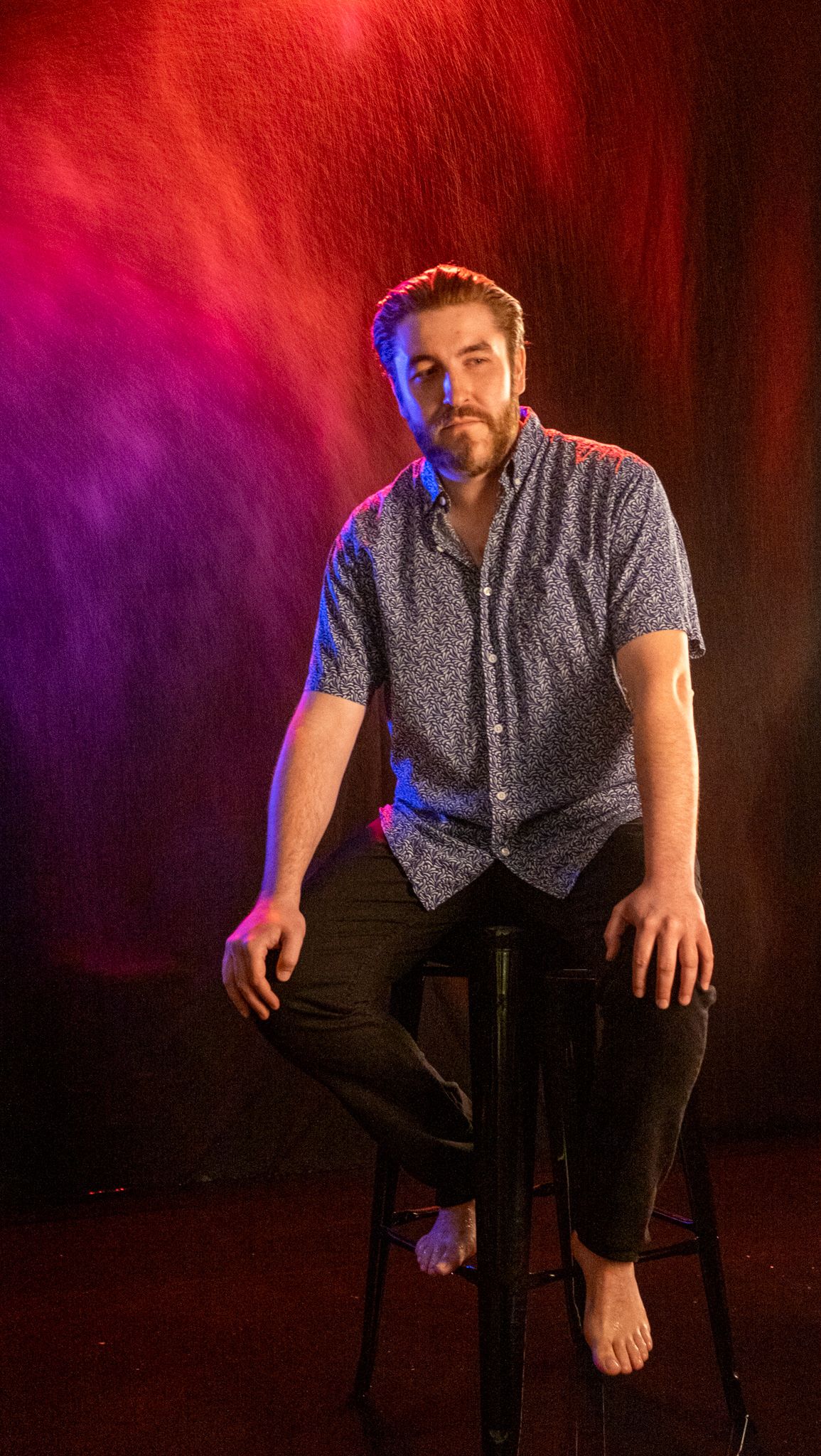 Bearded man in a patterned short-sleeve shirt and black pants sits barefoot on a stool against a red and purple lit backdrop.