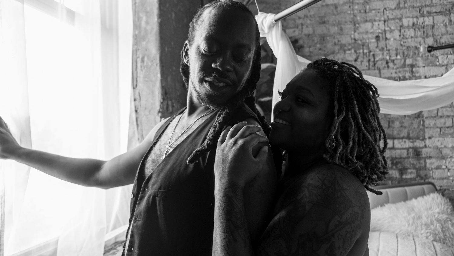 Black and white photo of two people embracing near a sunlit window beside an exposed brick wall.